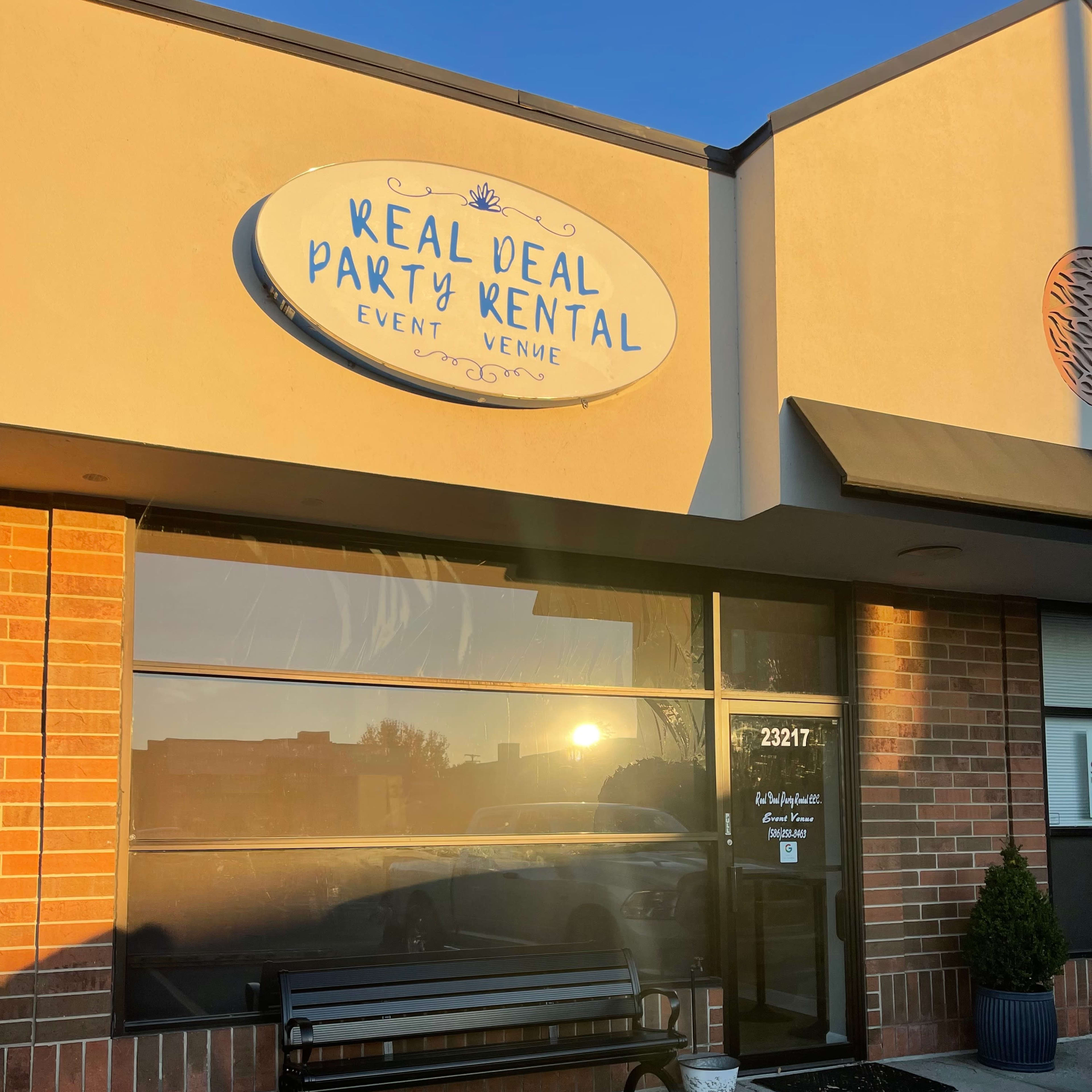 The exterior of a storefront features a sign that reads "Real Deal Party Rental Event Venue" above large windows reflecting the sunlight.