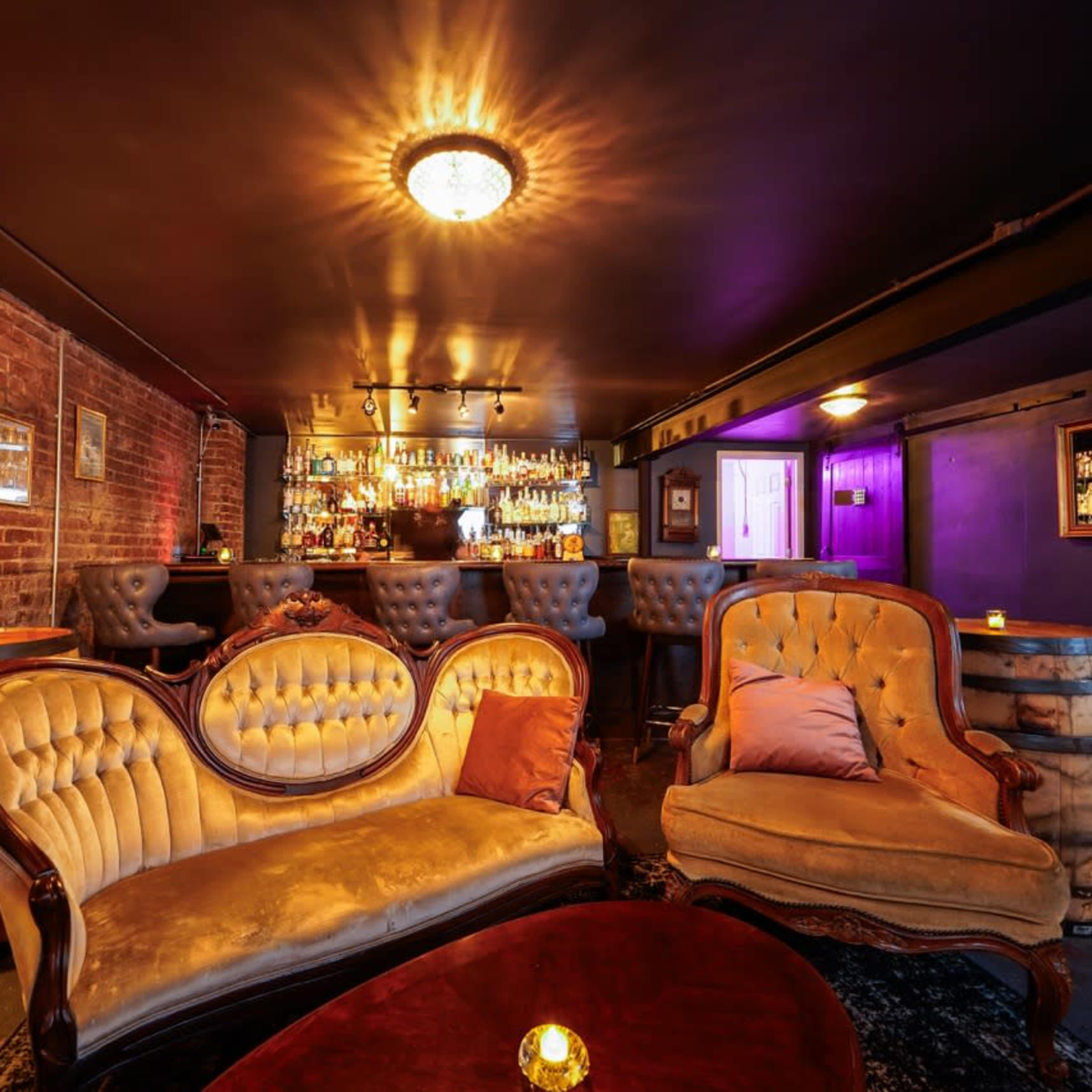 A dimly lit bar with a vintage seating area featuring a tufted couch and armchairs, and a bar counter lined with various bottles in the background.