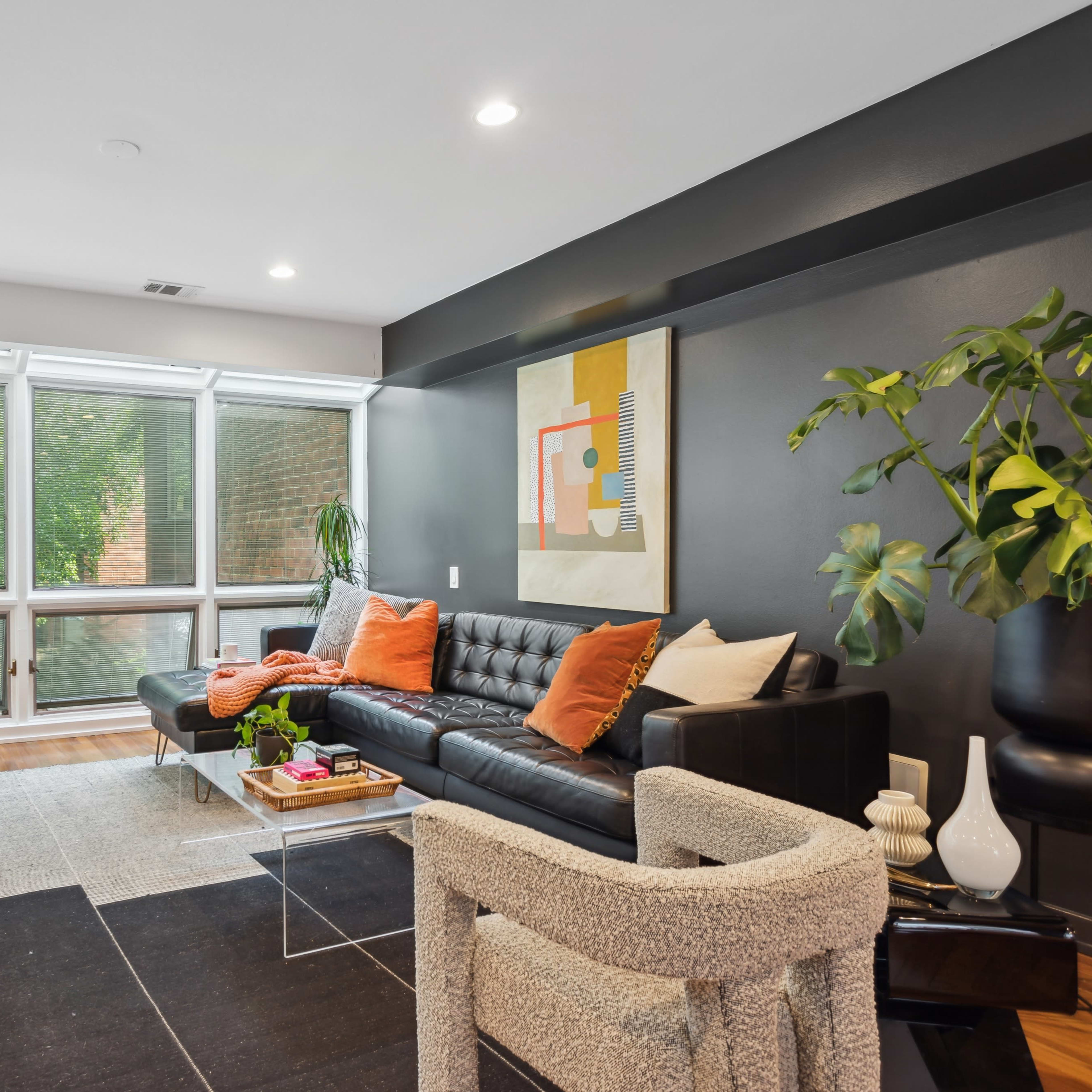 A modern living room with large windows, a black leather sofa, and a mix of decorative plants and furniture.