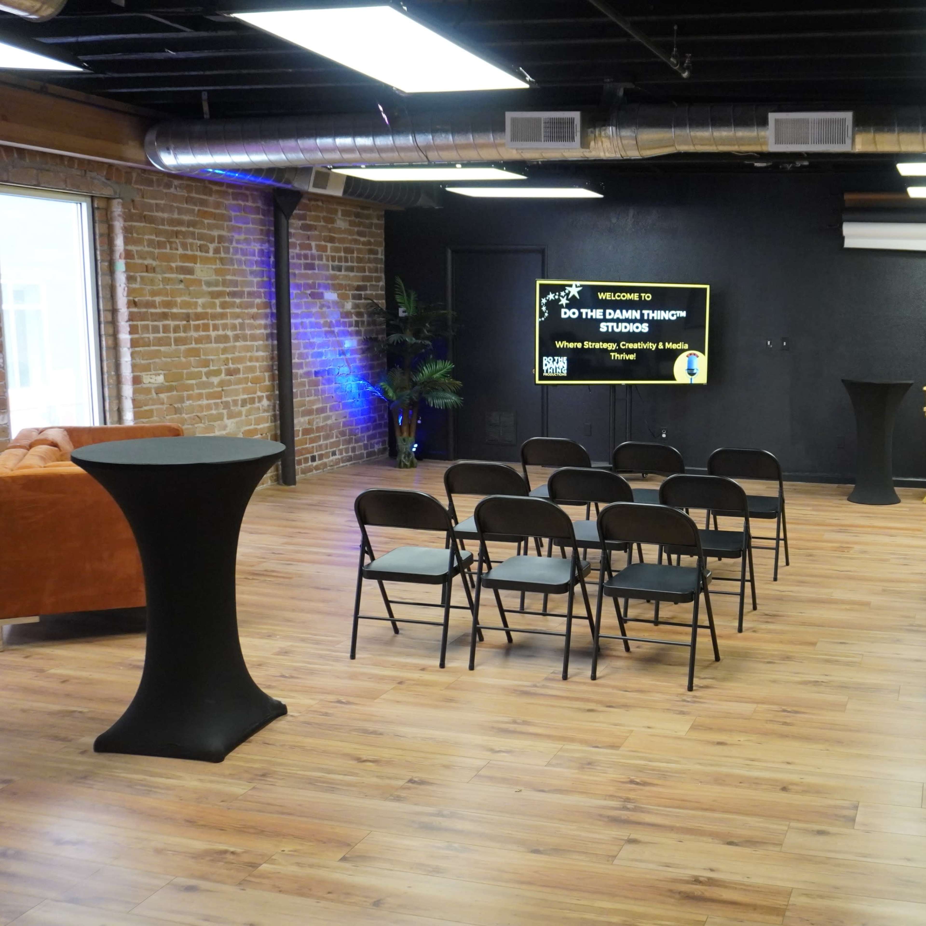 A neatly arranged event space with black chairs facing a screen, alongside a small podium and a couch against a brick wall.