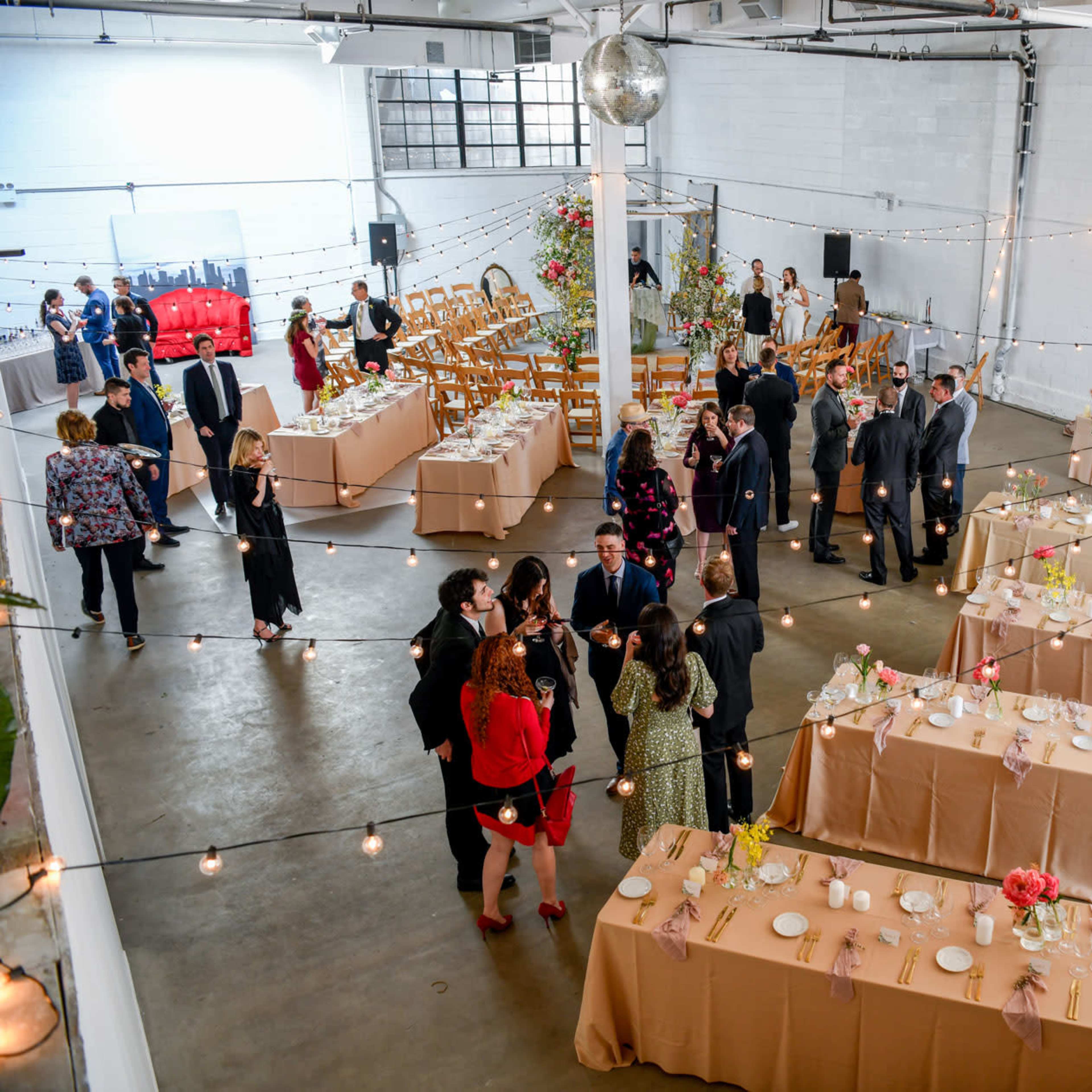 A spacious event venue features decorated tables, guests mingling, and string lights overhead.