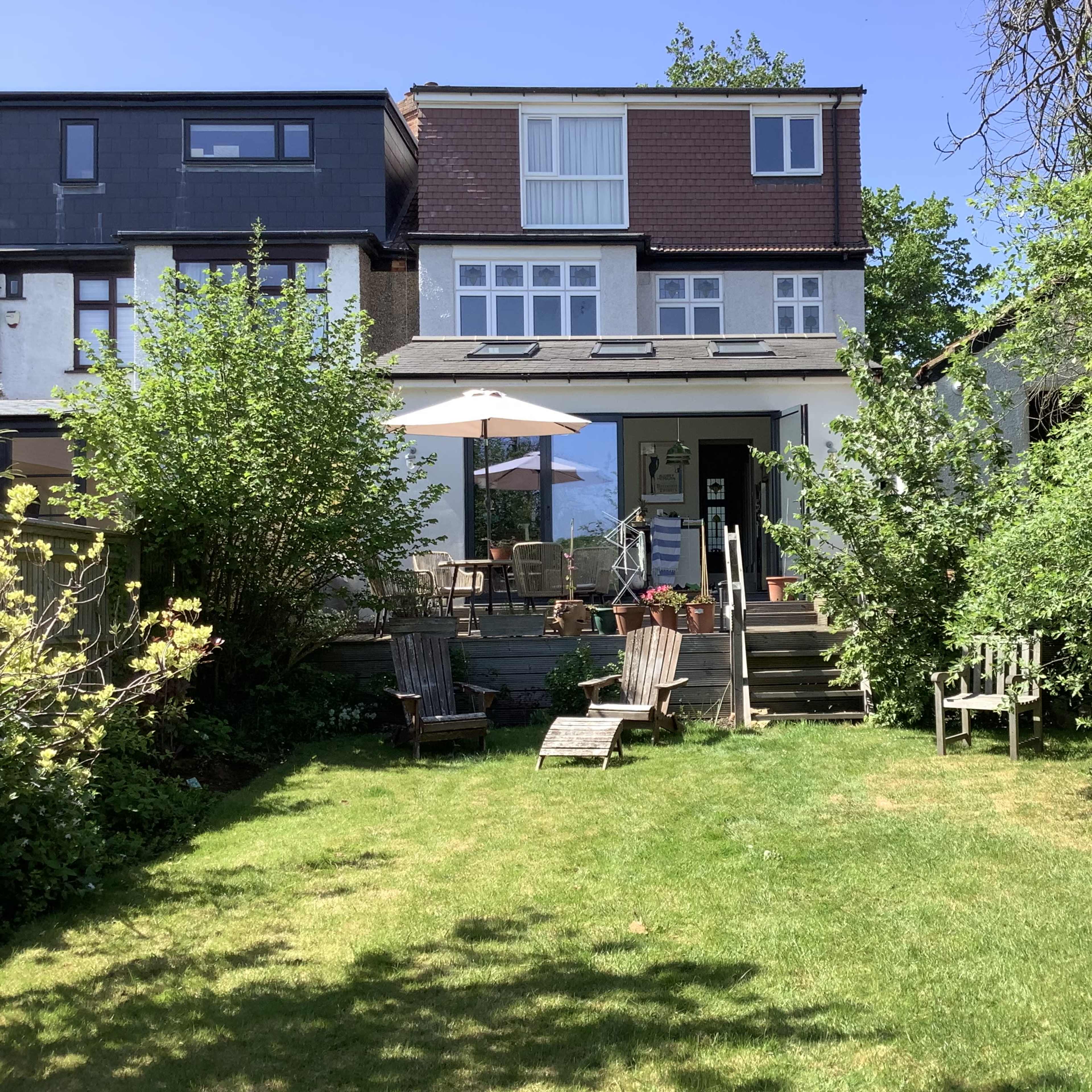 A two-story house with a patio sits at the back of a green backyard, surrounded by trees and lawn chairs.