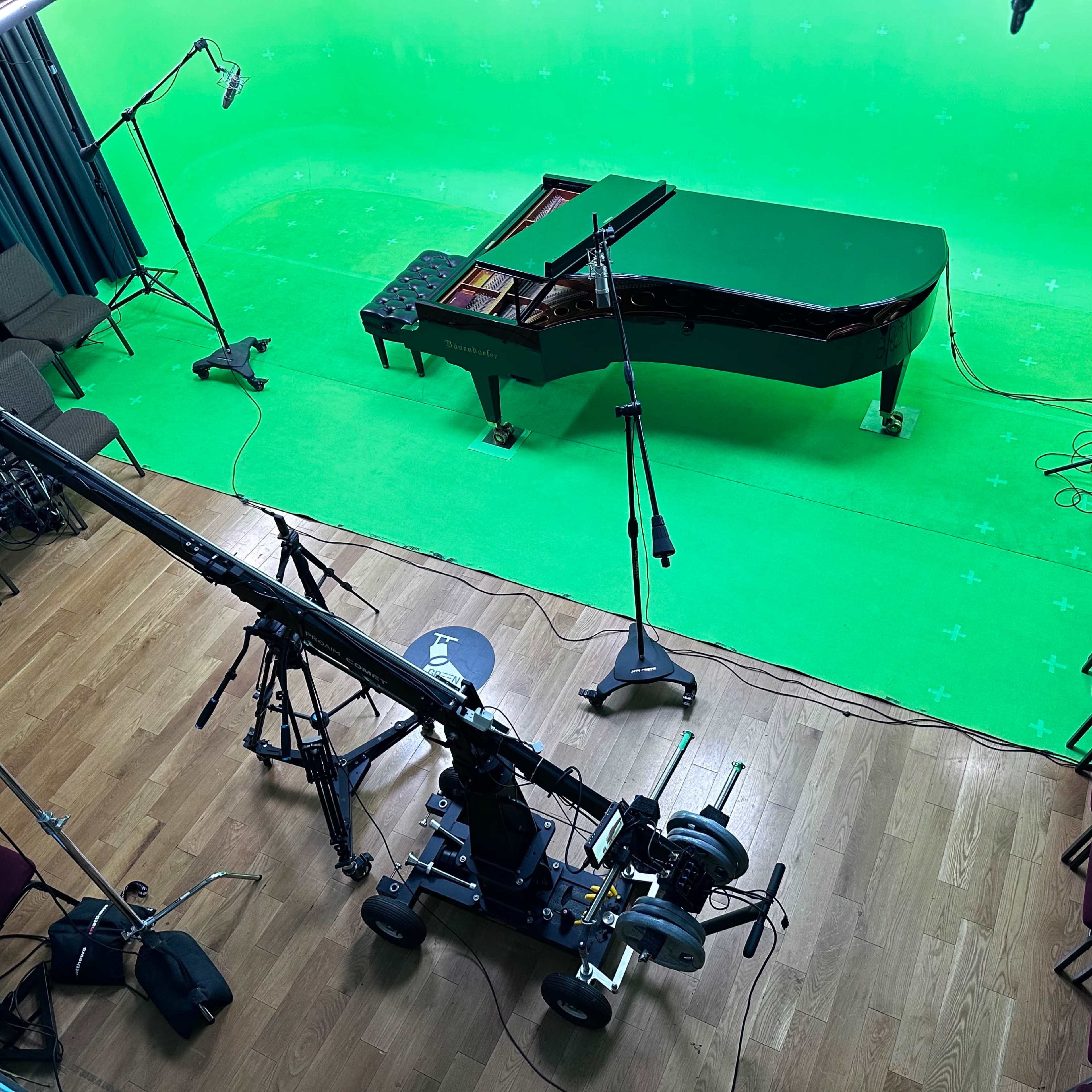 A grand piano is positioned on a green screen stage surrounded by lighting equipment and a camera setup.