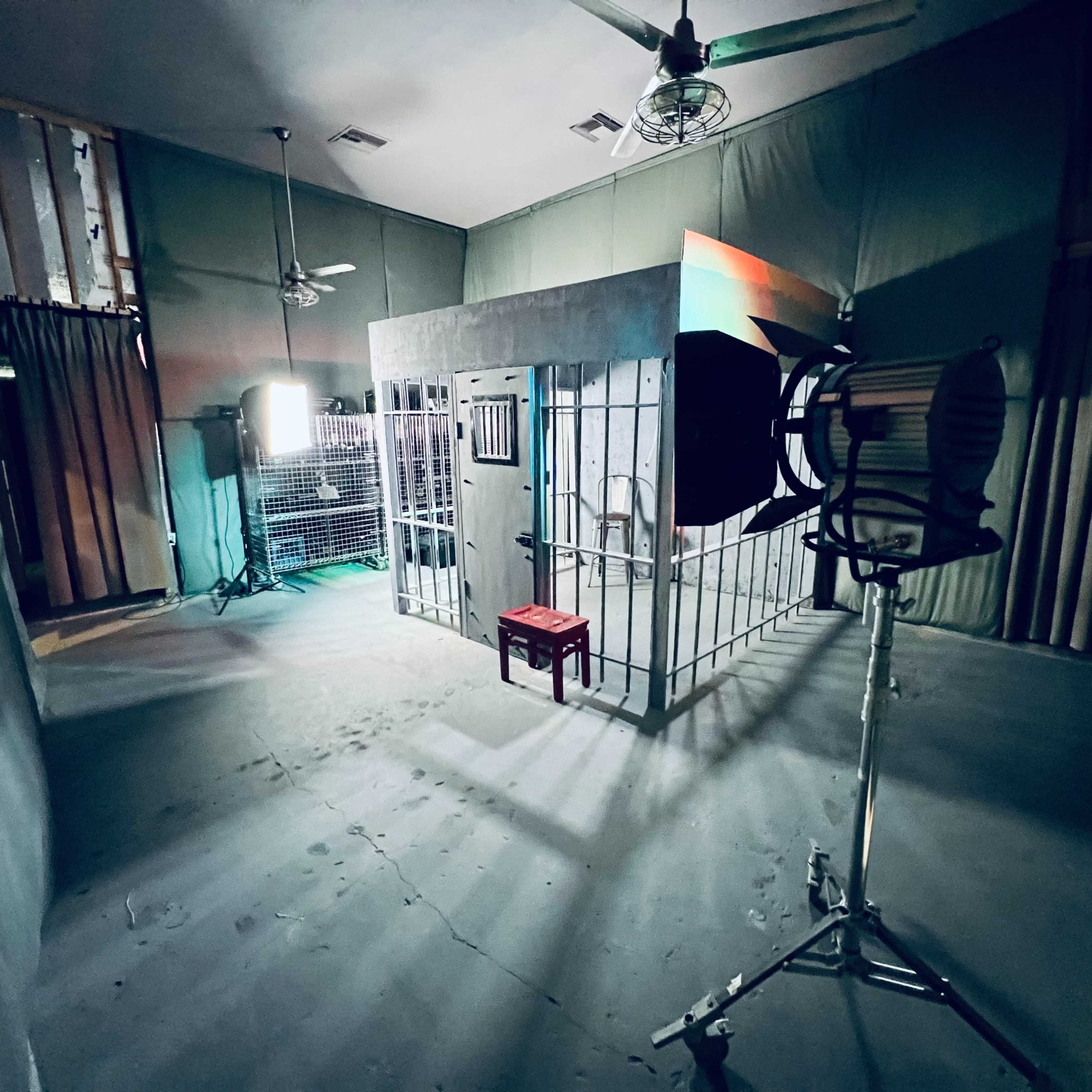A dimly lit room featuring a jail cell with metal bars, a red stool in front, and studio lighting equipment set up nearby.