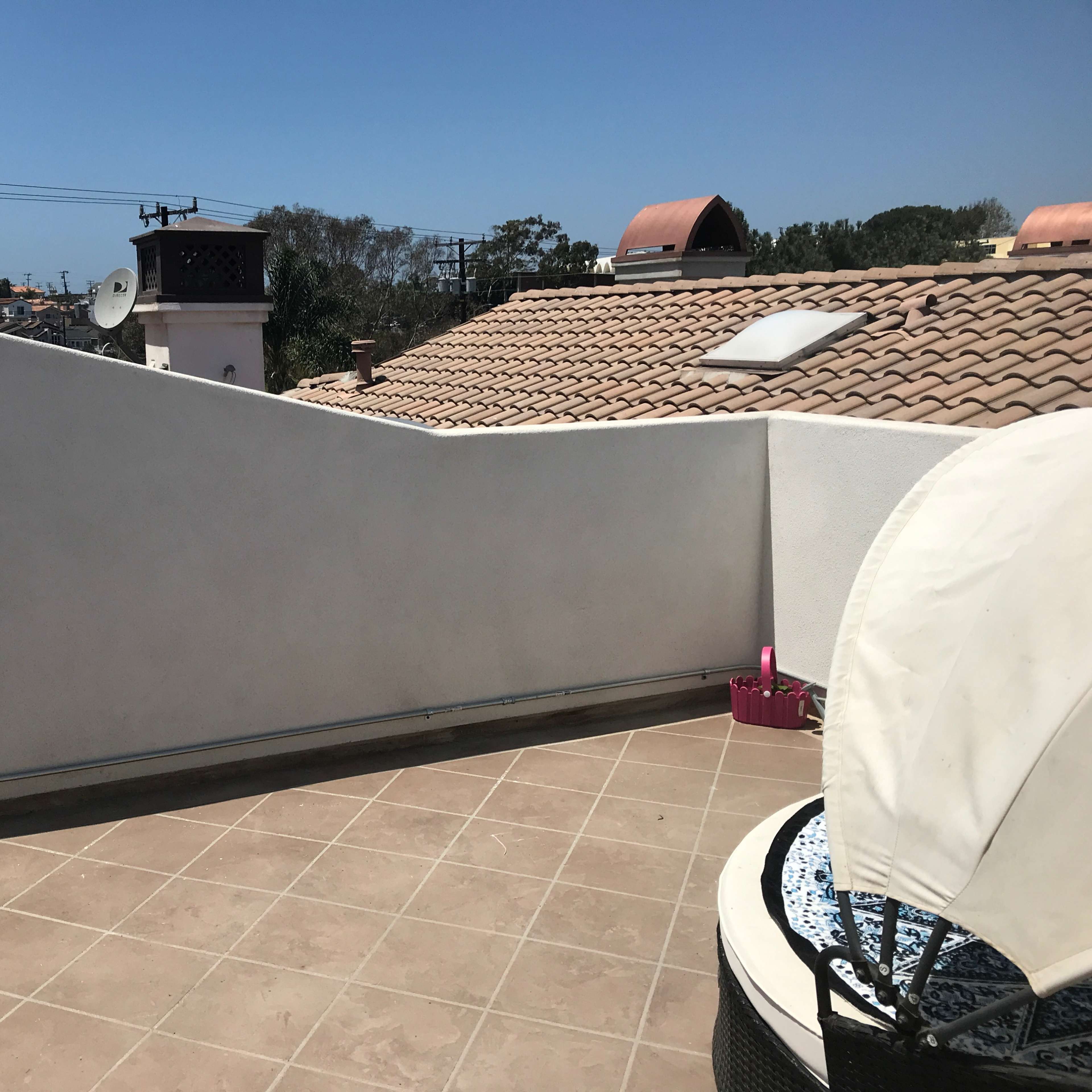 A rooftop terrace with a tiled floor, a curved white wall, and a view of neighboring rooftops under a clear blue sky.