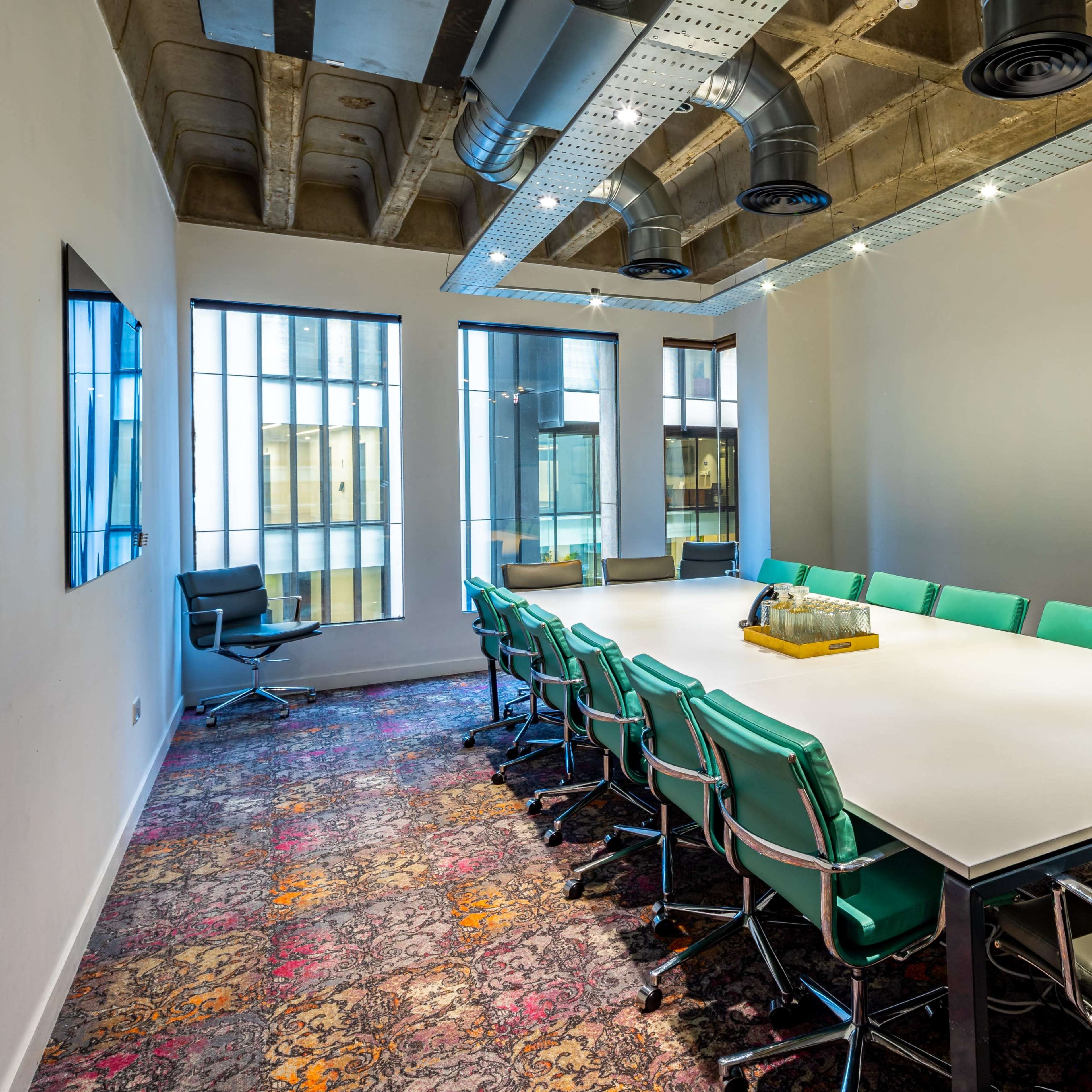 A large conference room features a rectangular table surrounded by turquoise and silver chairs, with large windows and exposed ceiling elements.