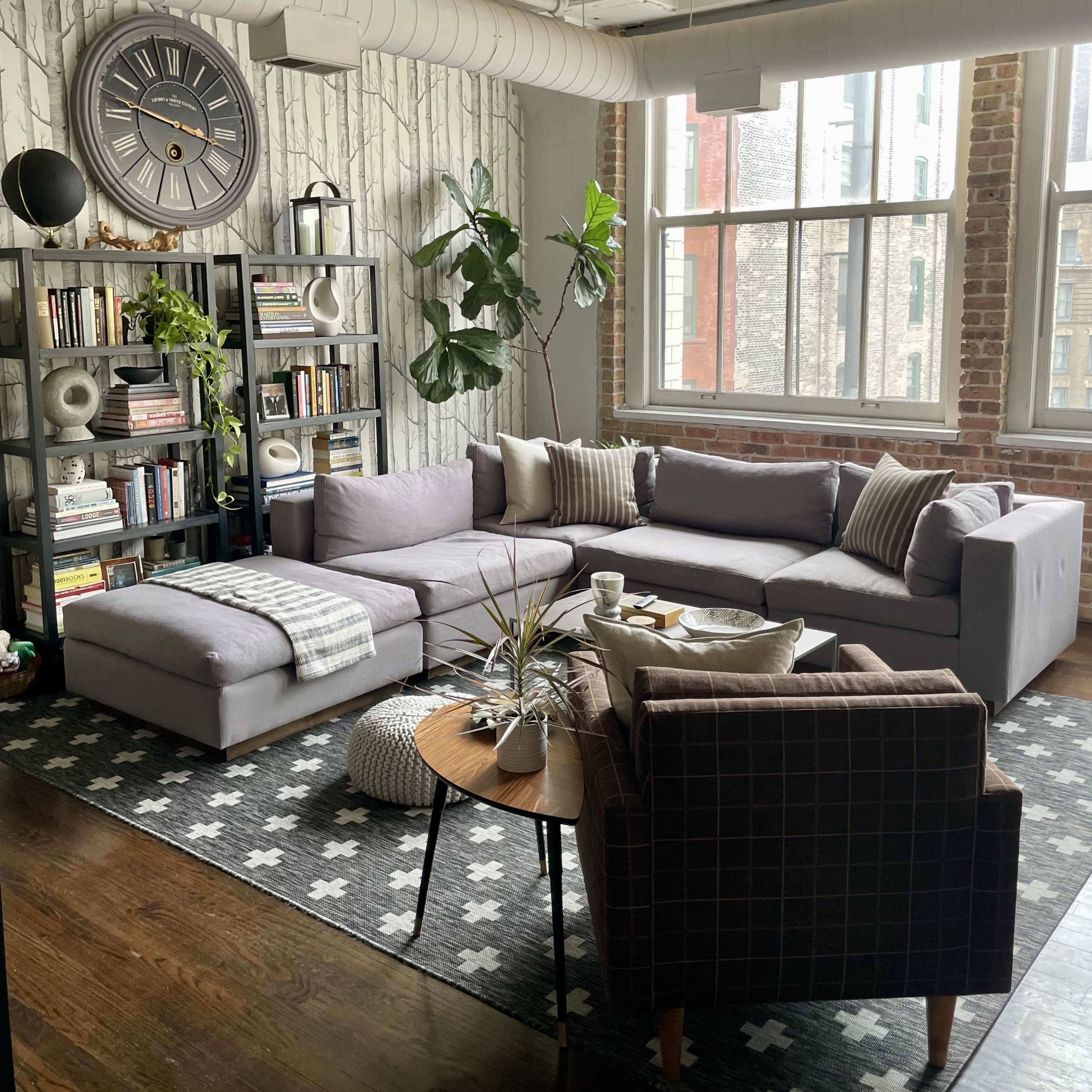 A modern living room features a gray sectional sofa, a round wooden coffee table, and a bookshelf filled with books, all set against a backdrop of brick walls and large windows.