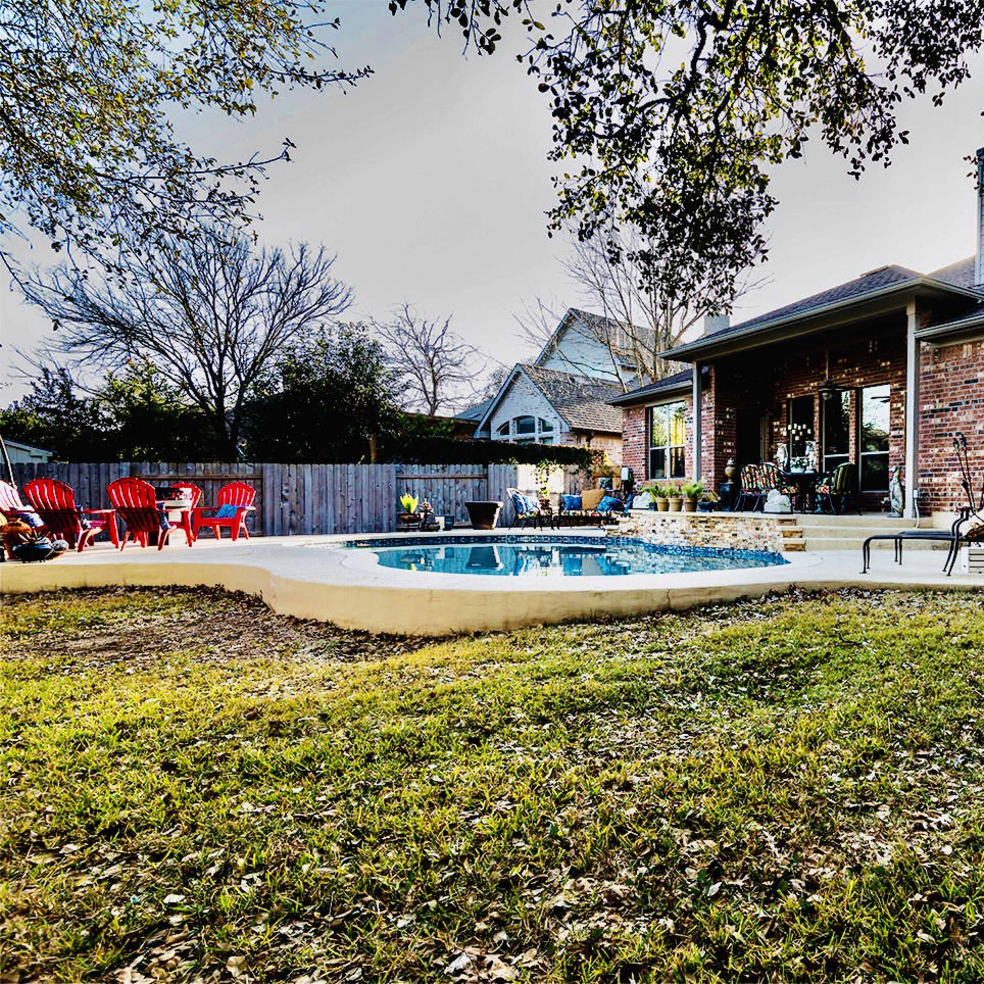 A backyard features a pool surrounded by lounge chairs and a seating area near a brick house.
