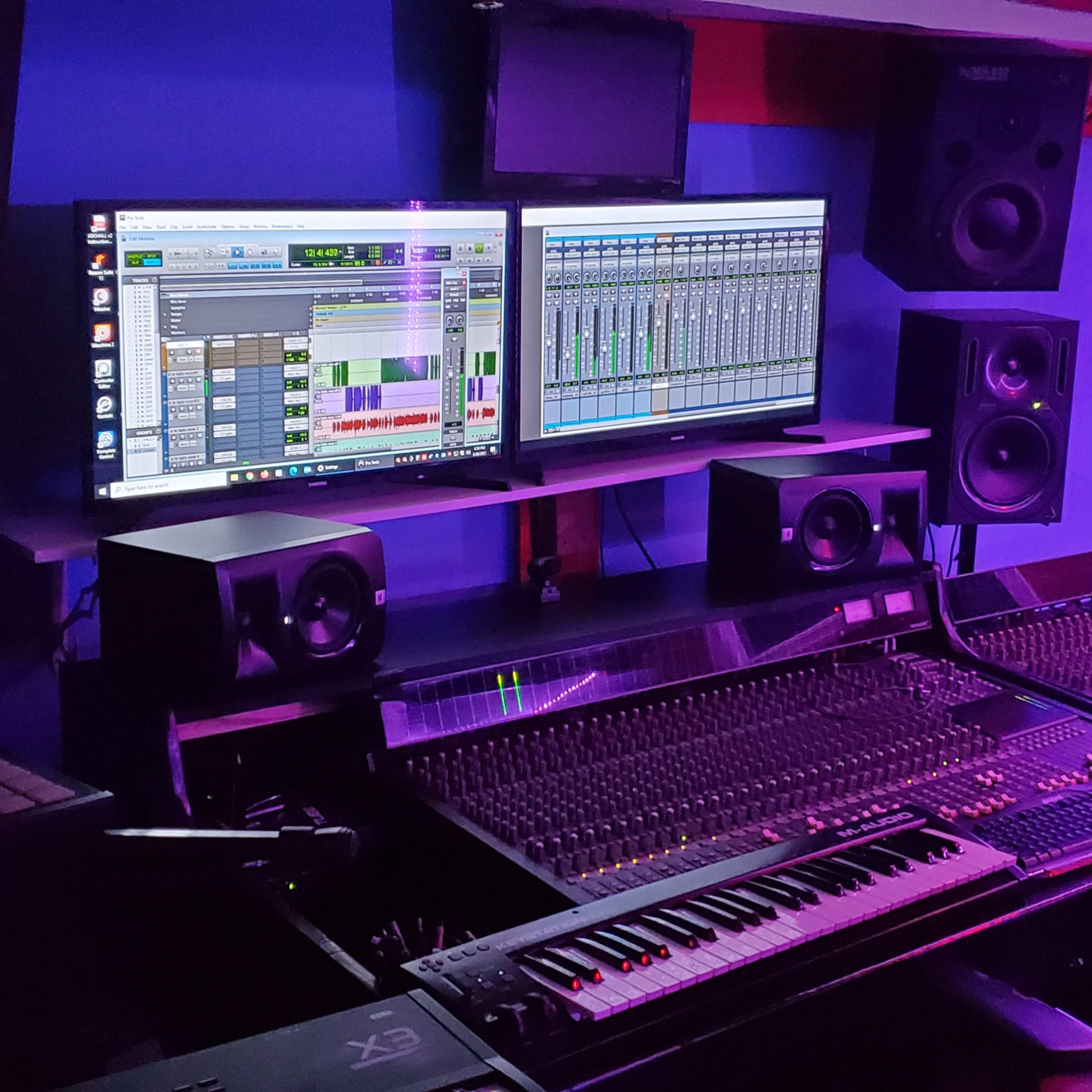 A music studio with a digital mixing console, two large computer monitors displaying audio software, and several speakers arranged around the setup.