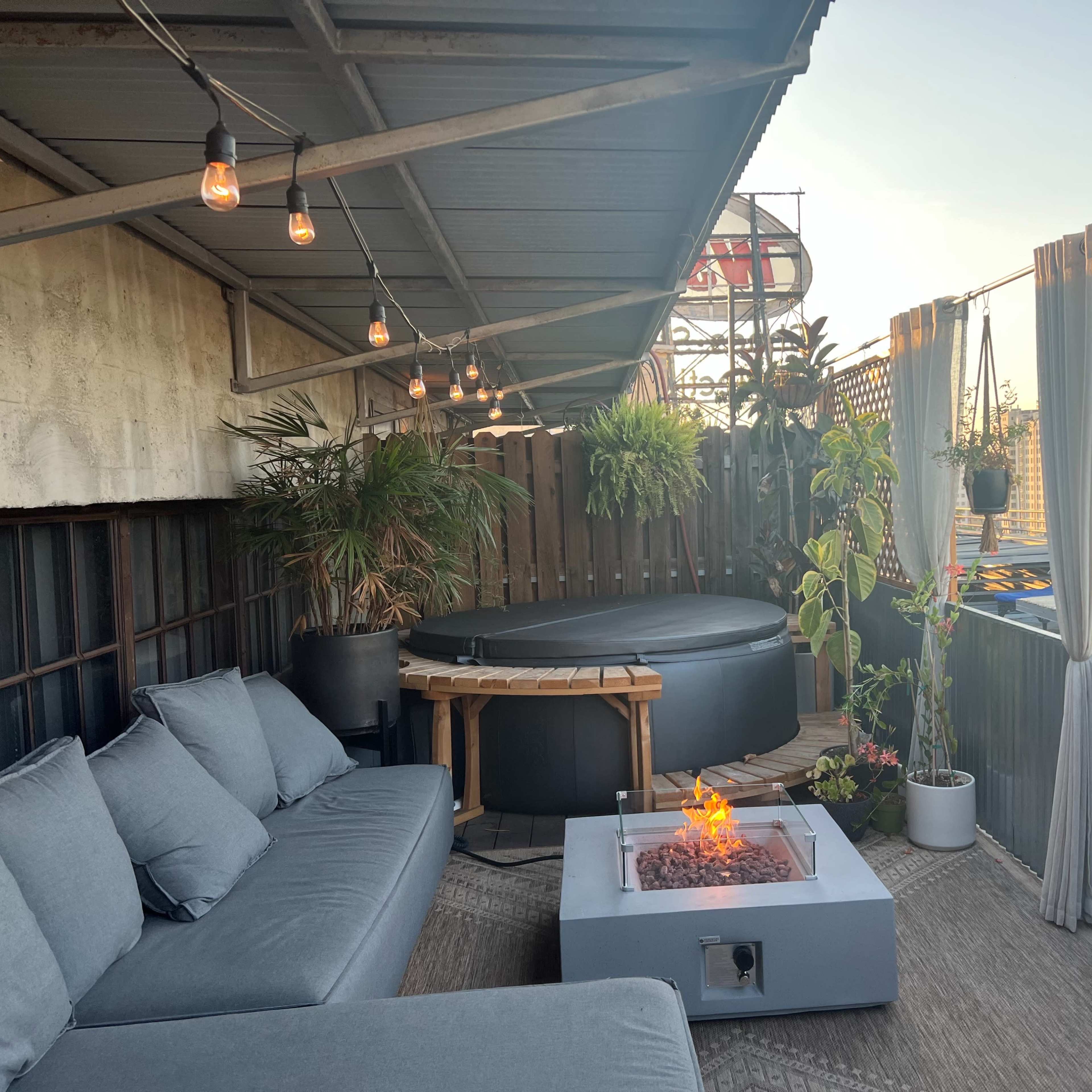 The scene features a cozy rooftop space with a hot tub, comfortable seating, and potted plants, illuminated by string lights.