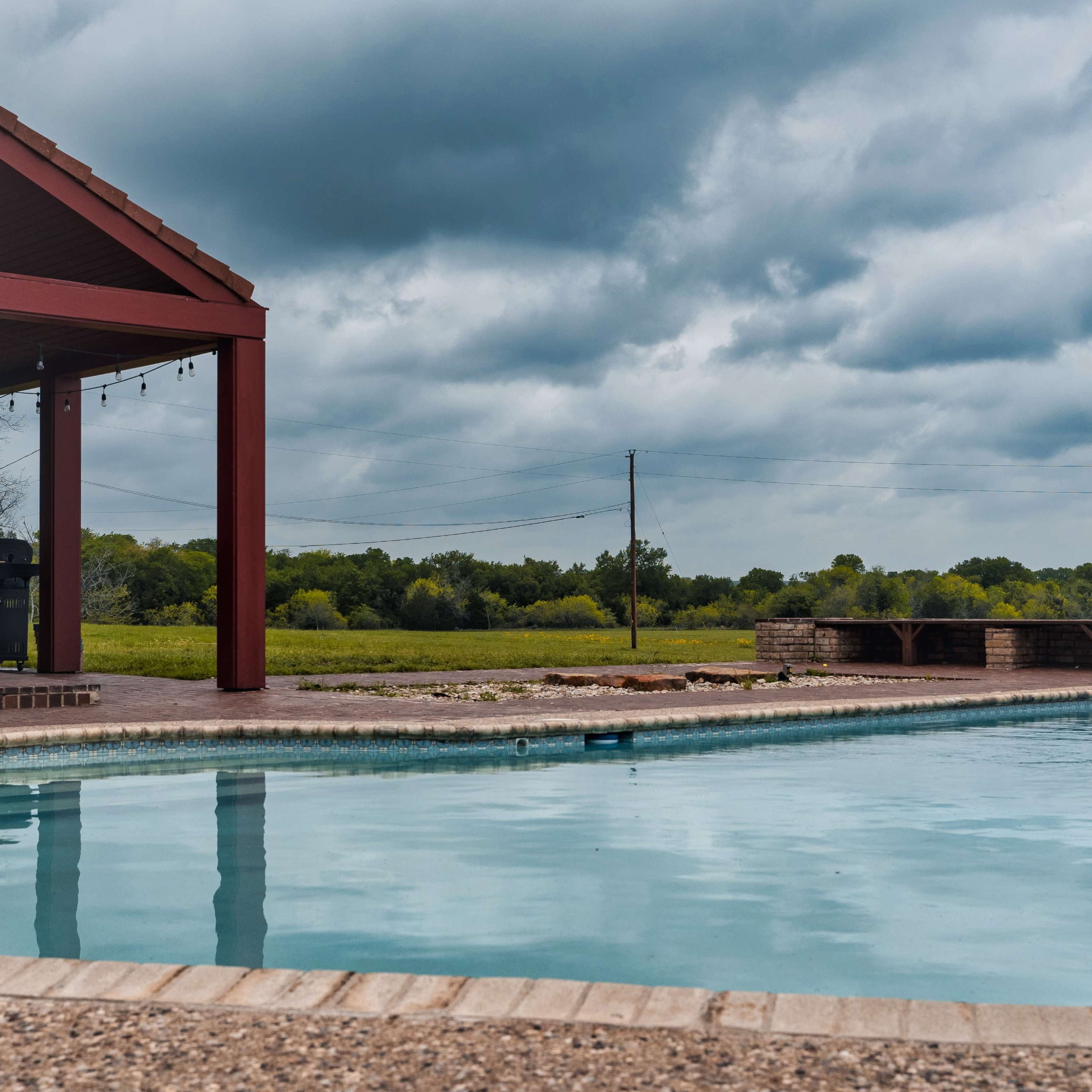 A swimming pool reflects the sky under a canopy of gray clouds, with a red-roofed patio and a grassy field in the background.