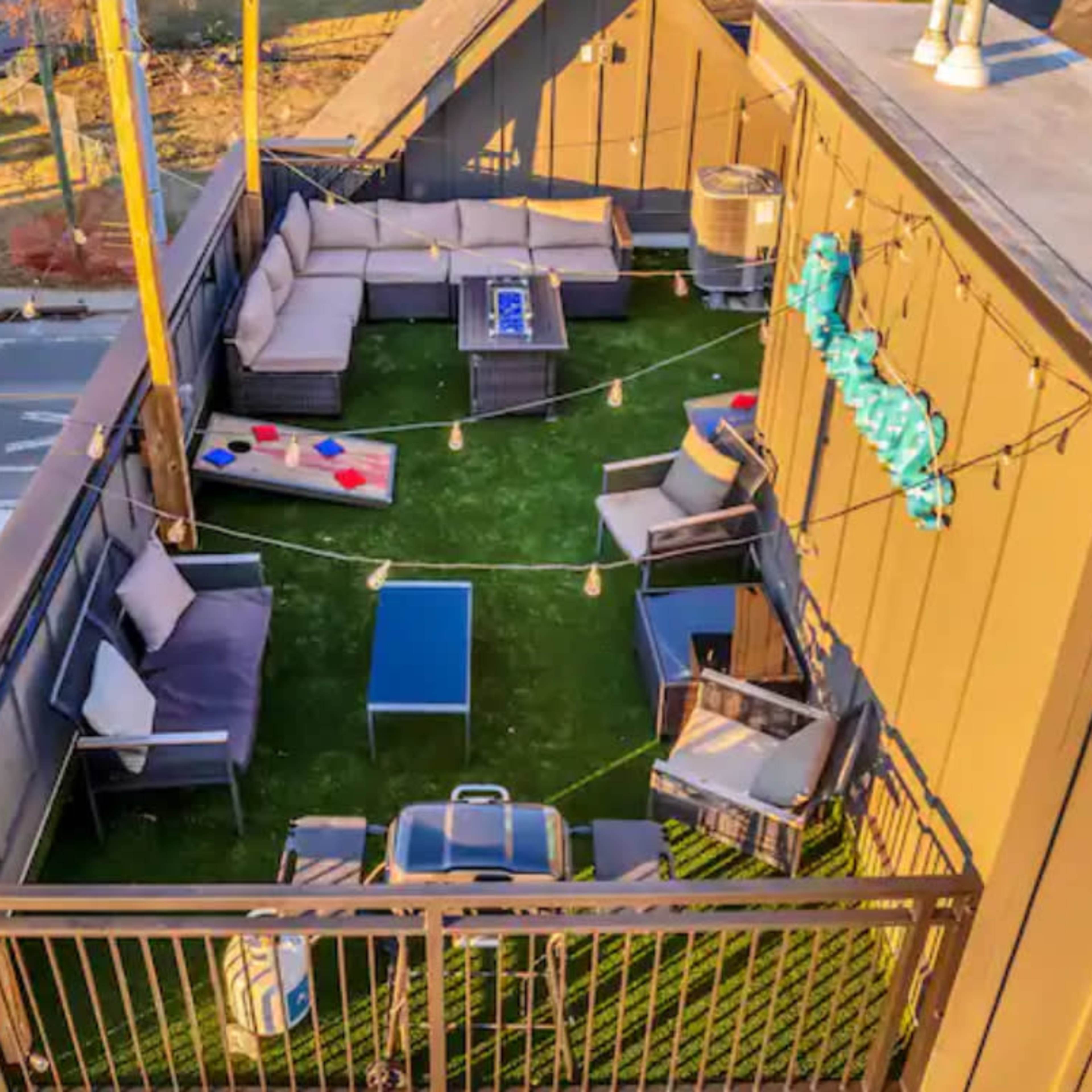 A rooftop terrace with green artificial turf, a seating area featuring gray couches and a blue coffee table, and a barbecue grill.