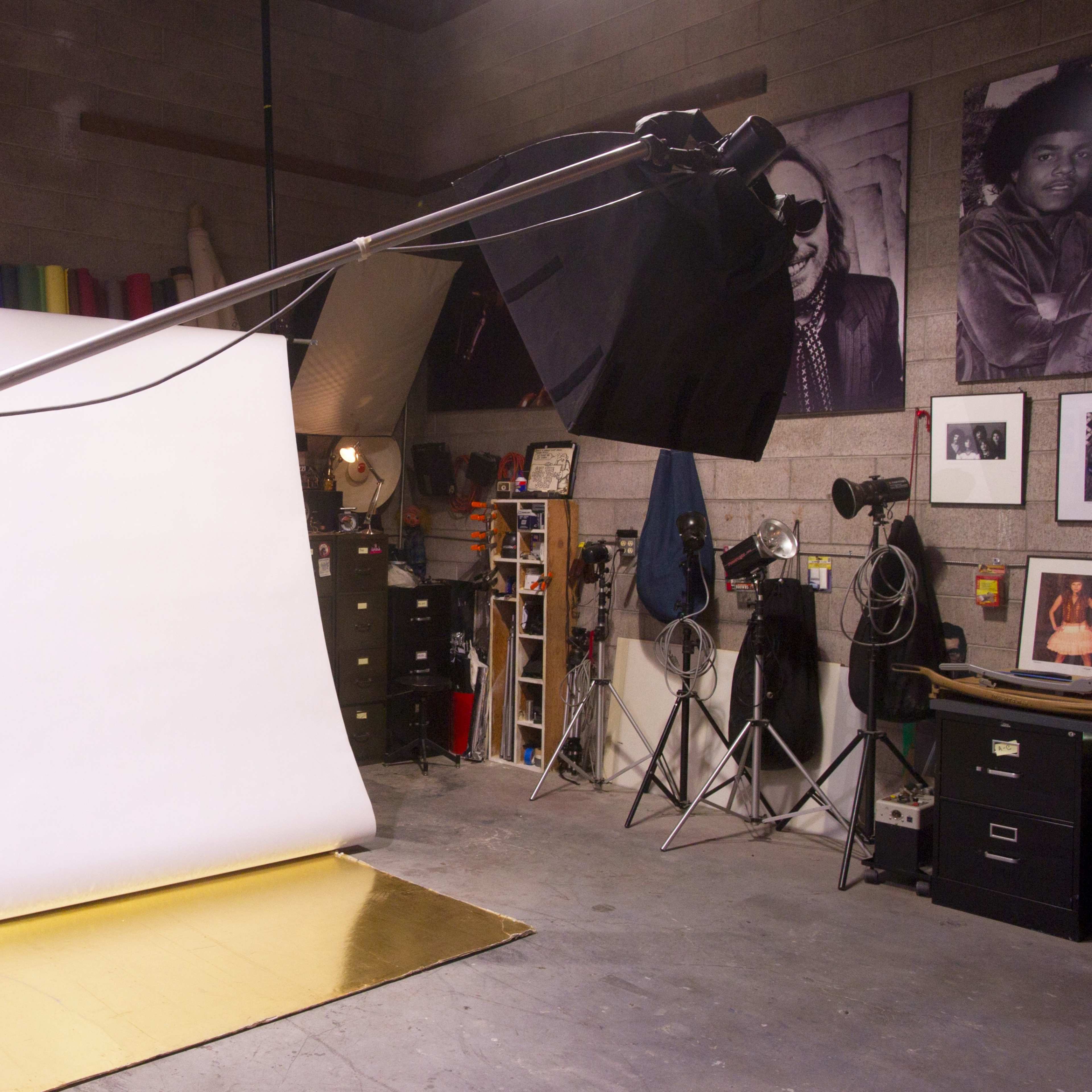 A photography studio features a white backdrop on a stand, a golden floor, and various lighting equipment, with a mannequin positioned nearby.