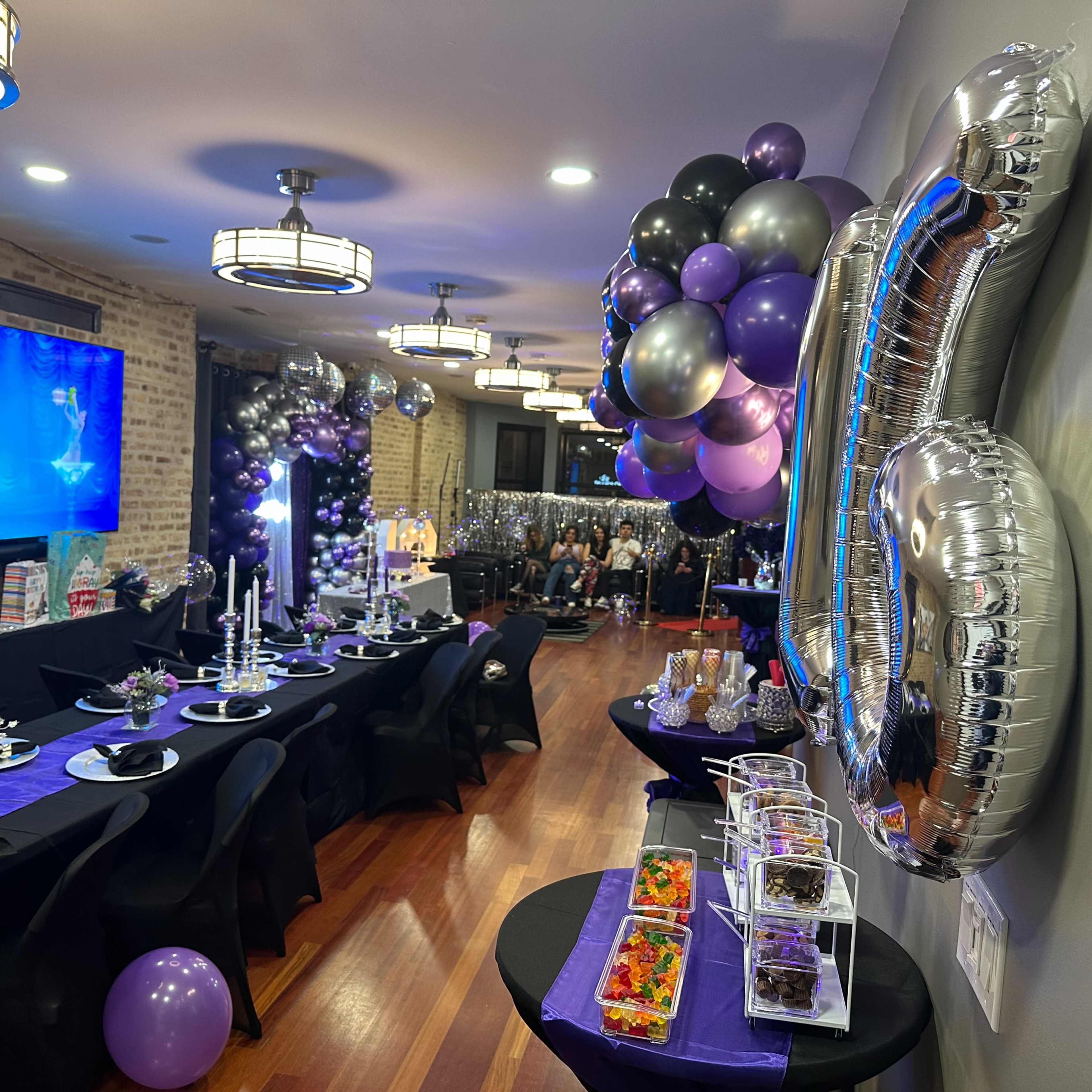 A decorated event space with a long table set for a party, featuring black and purple decor, appetizers, and a large balloon arch.