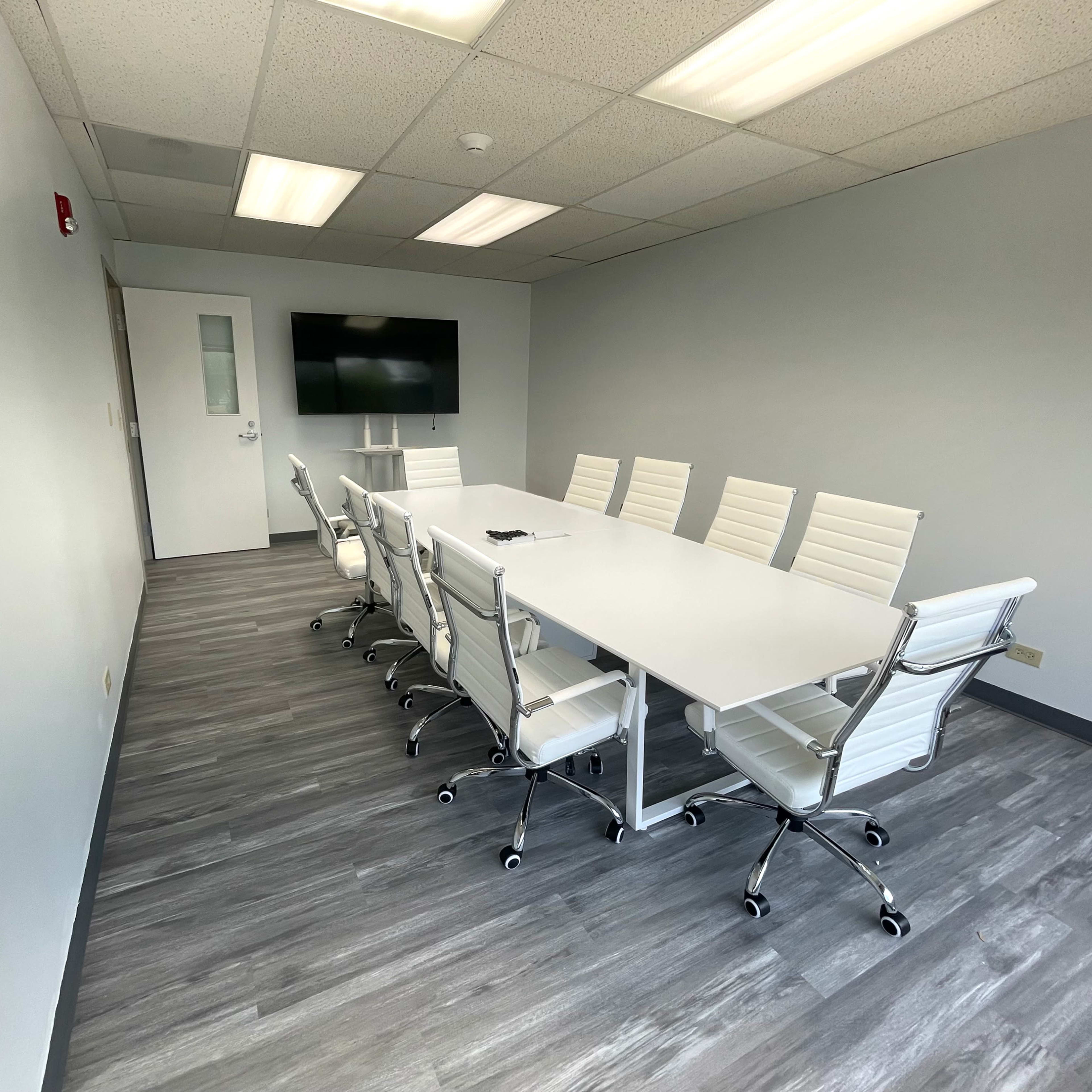 A sleek conference room features a long white table surrounded by eight white rolling chairs and a wall-mounted screen.