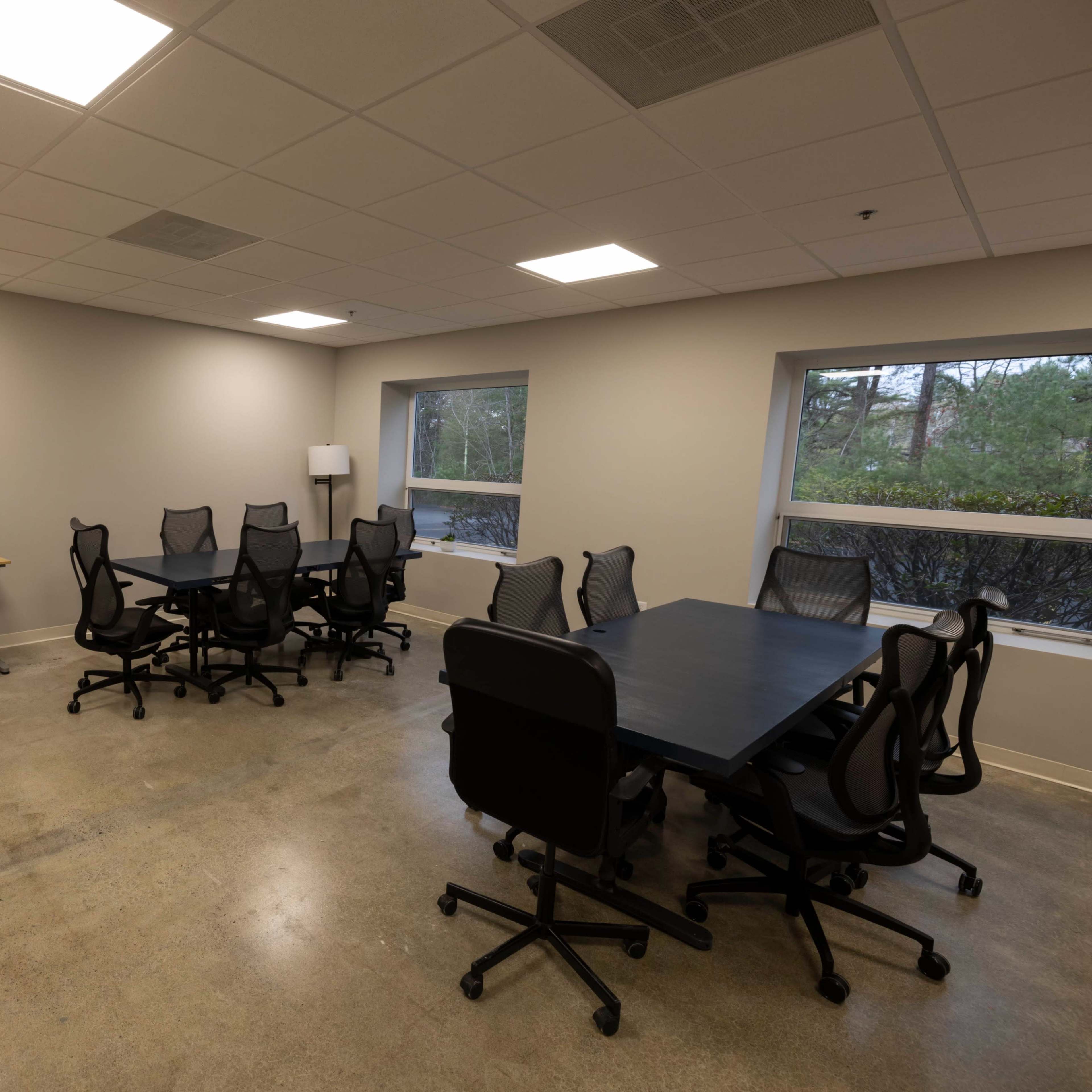 A well-lit meeting room with several black office chairs around dark tables, a printer on a desk, and large windows overlooking greenery.
