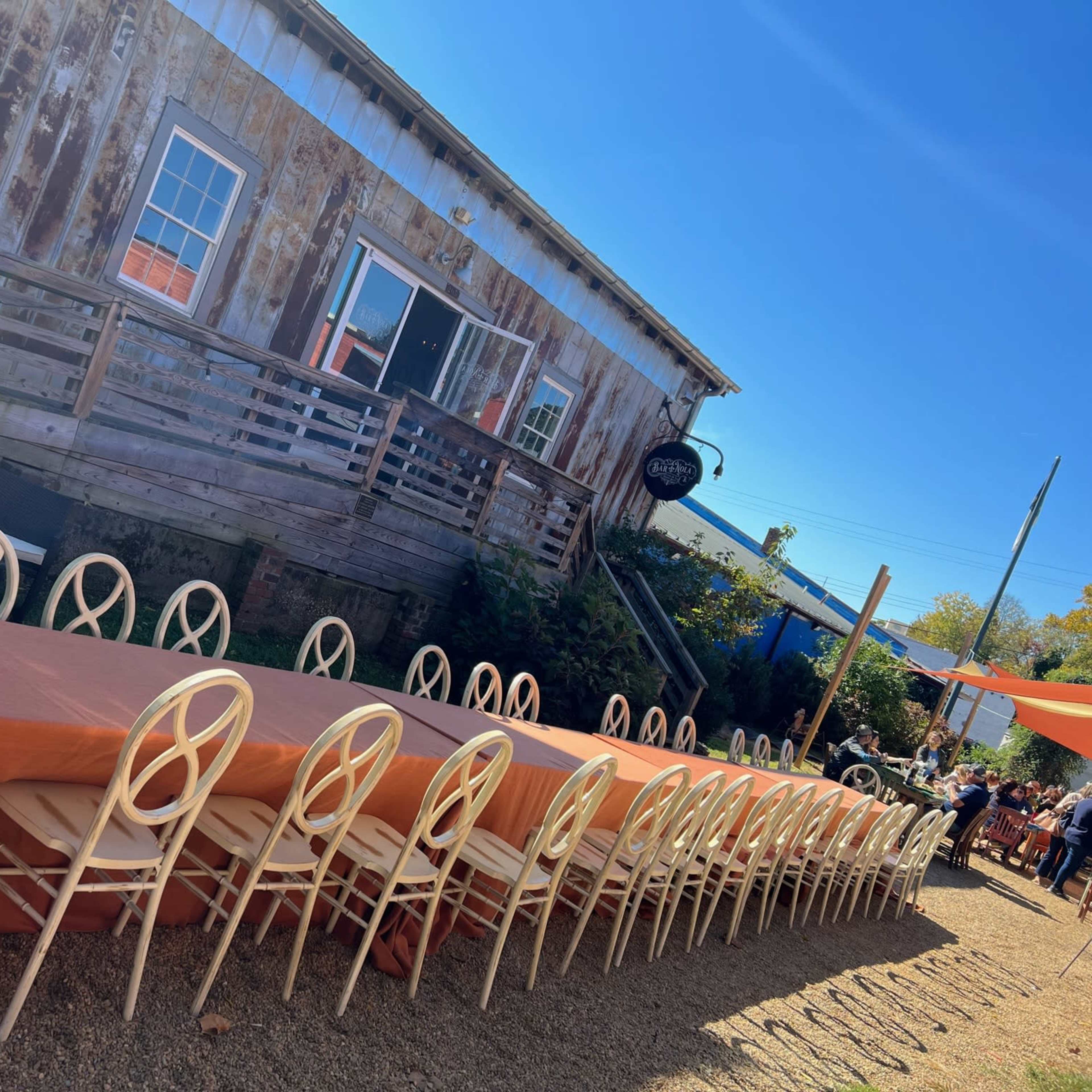 A long, rectangular table with an orange tablecloth is set up outdoors in front of a rustic building, surrounded by chairs.