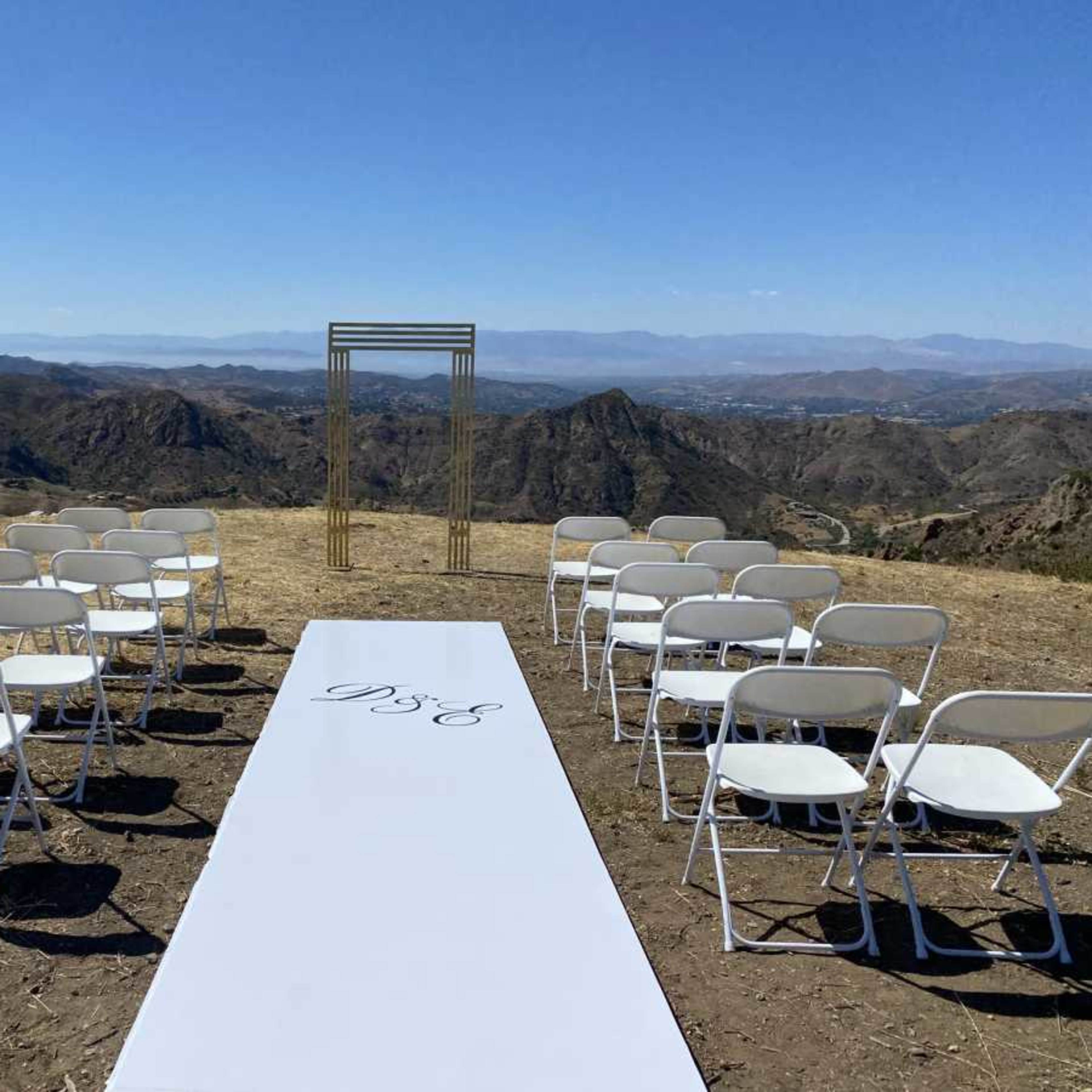 An outdoor wedding setup features rows of white chairs facing a decorated arch on a grassy area with a mountainous landscape in the background.