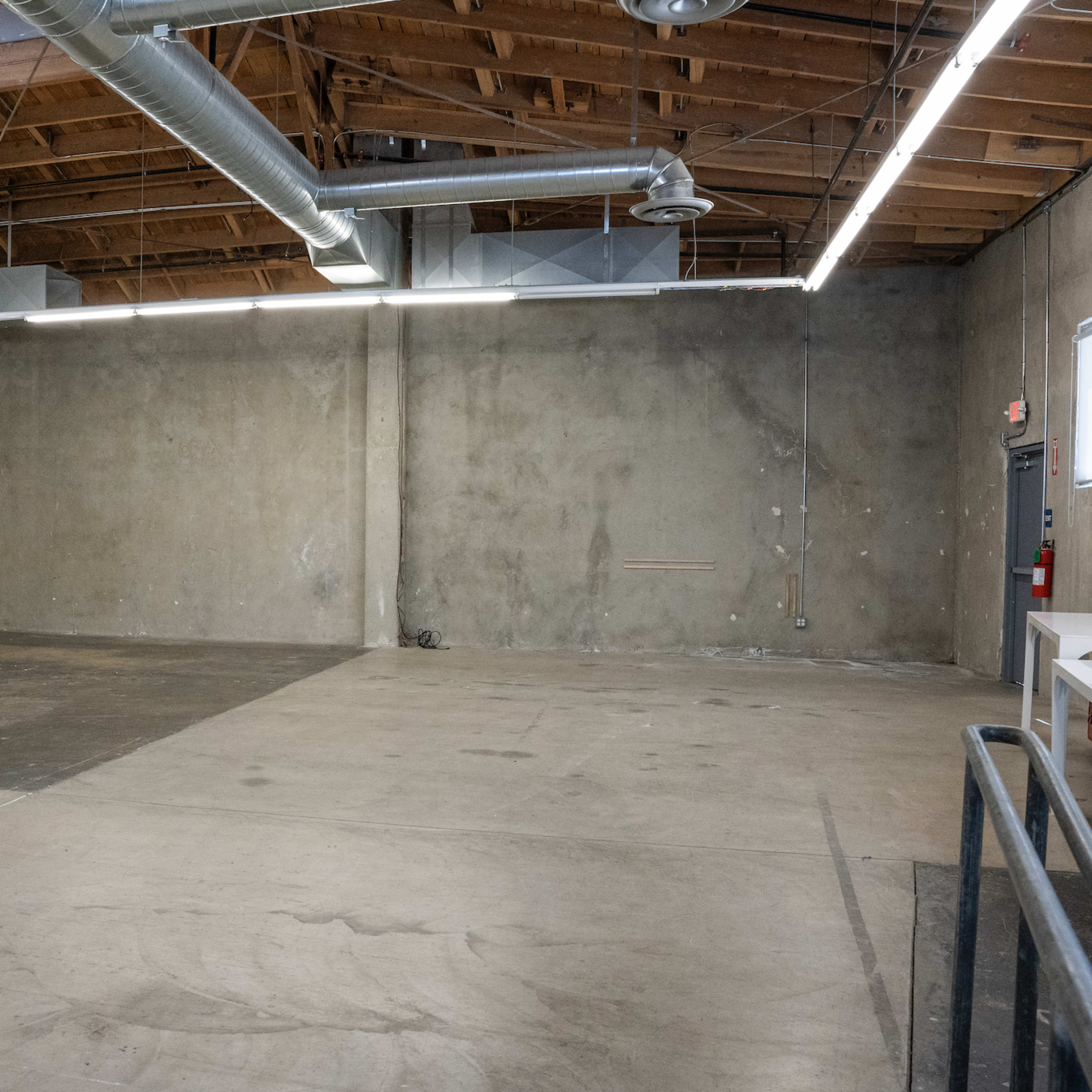 A spacious, industrial-style room with concrete floors, exposed wooden beams, and ductwork overhead, featuring minimal furnishings on one side.