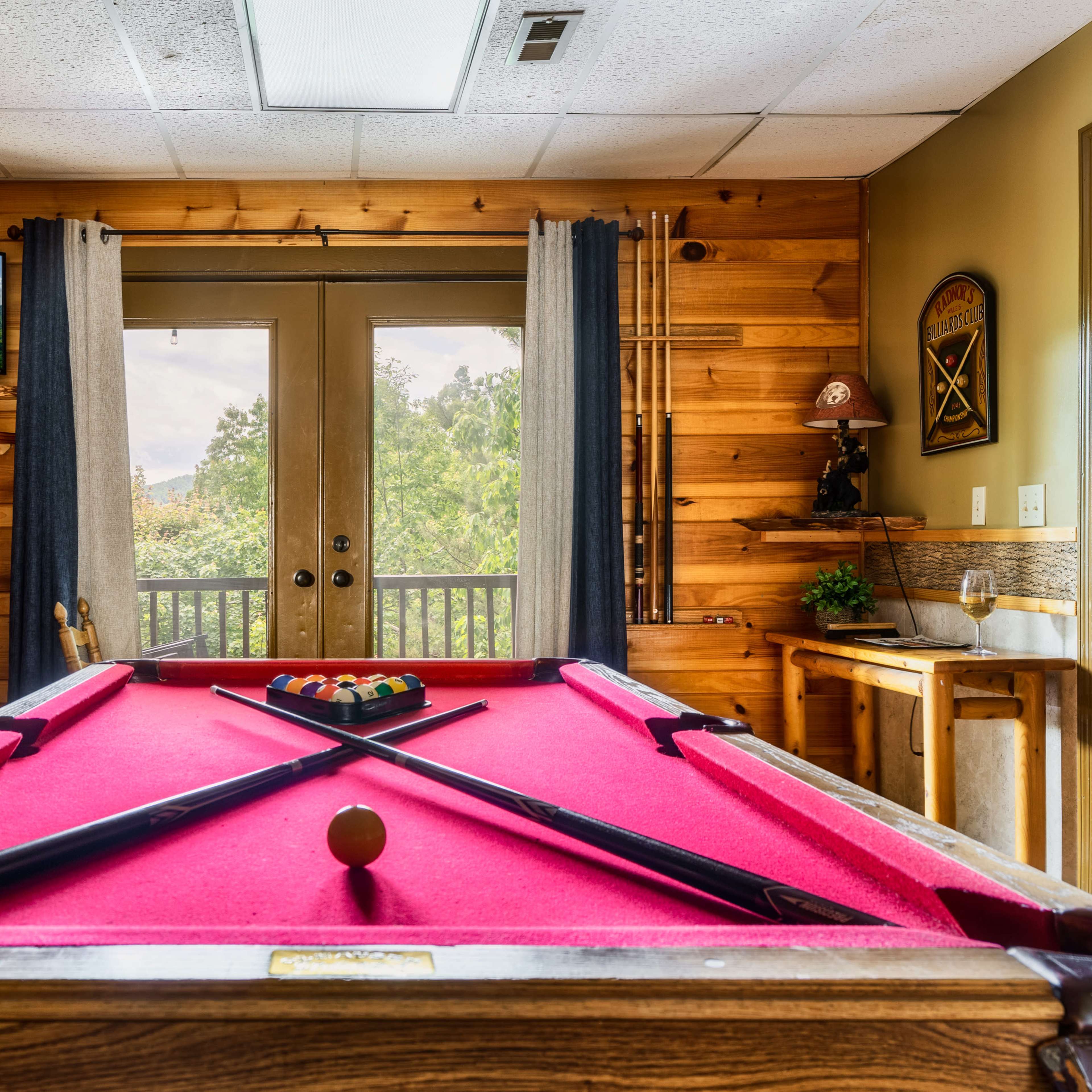 A pool table with a pink felt surface in a room with wooden walls and a view of trees through the double doors.