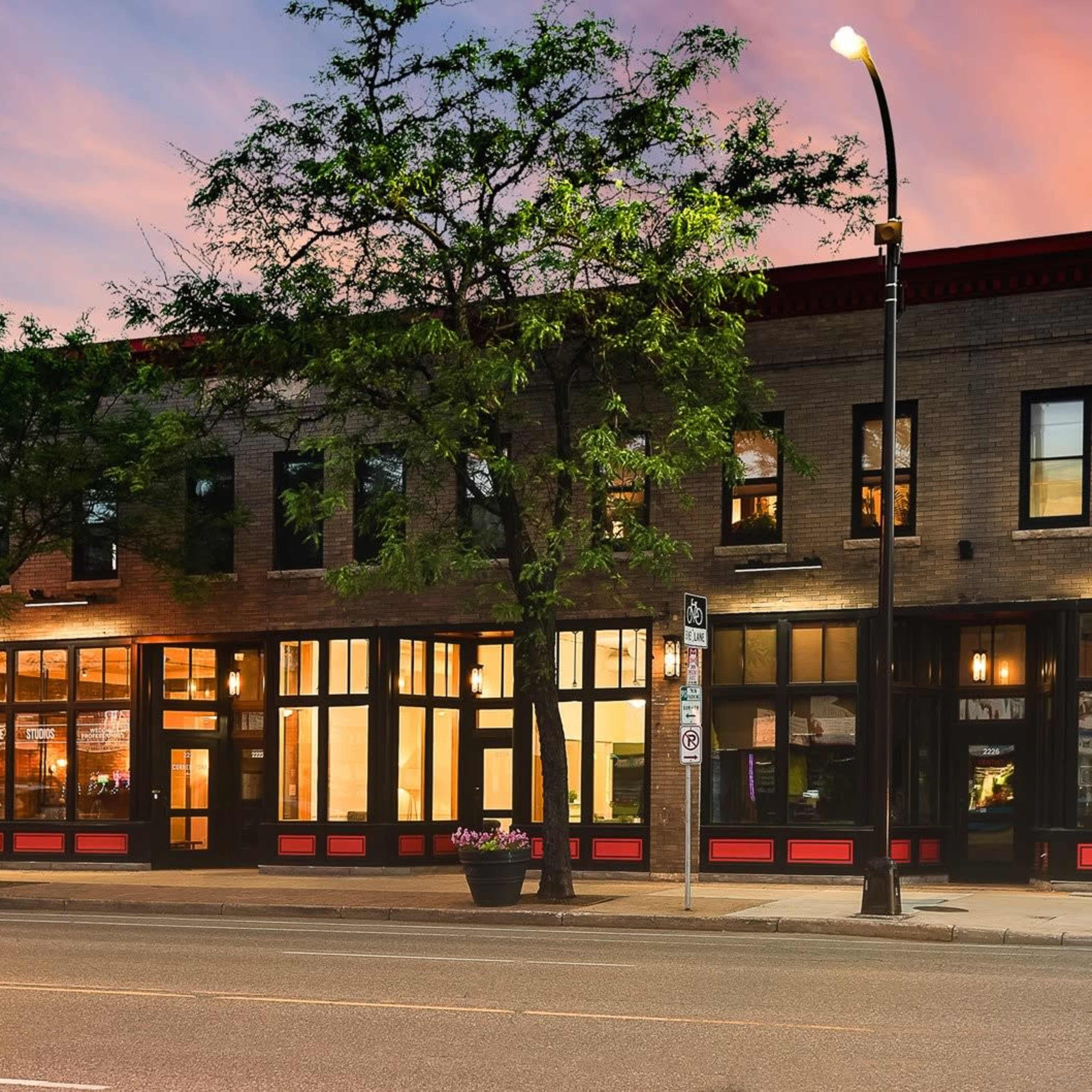 A two-story brick building with large windows, located on a city street at dusk, featuring a red and black color scheme.