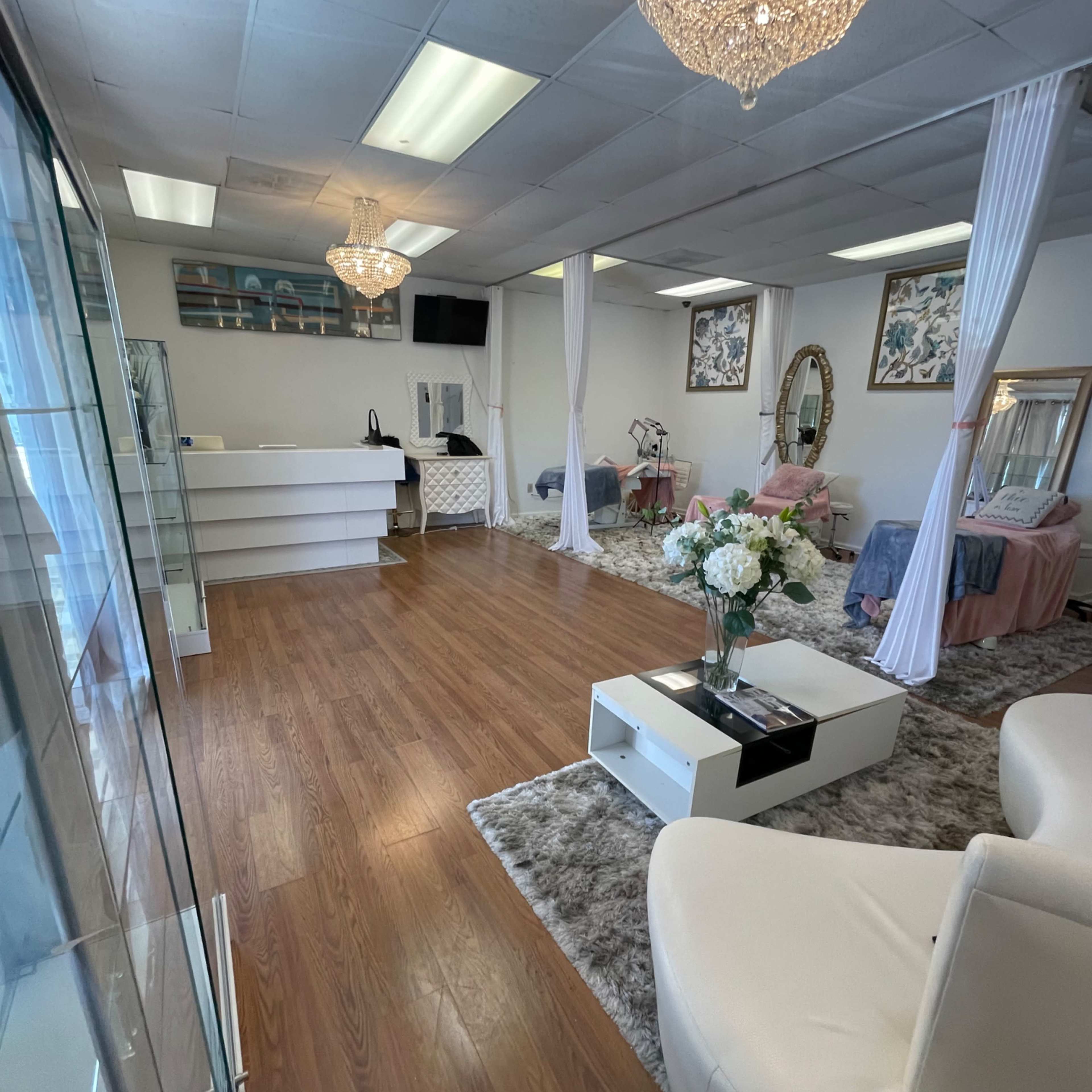A spacious, well-lit salon interior featuring white furniture, decorative chandeliers, and treatment areas with draped curtains.