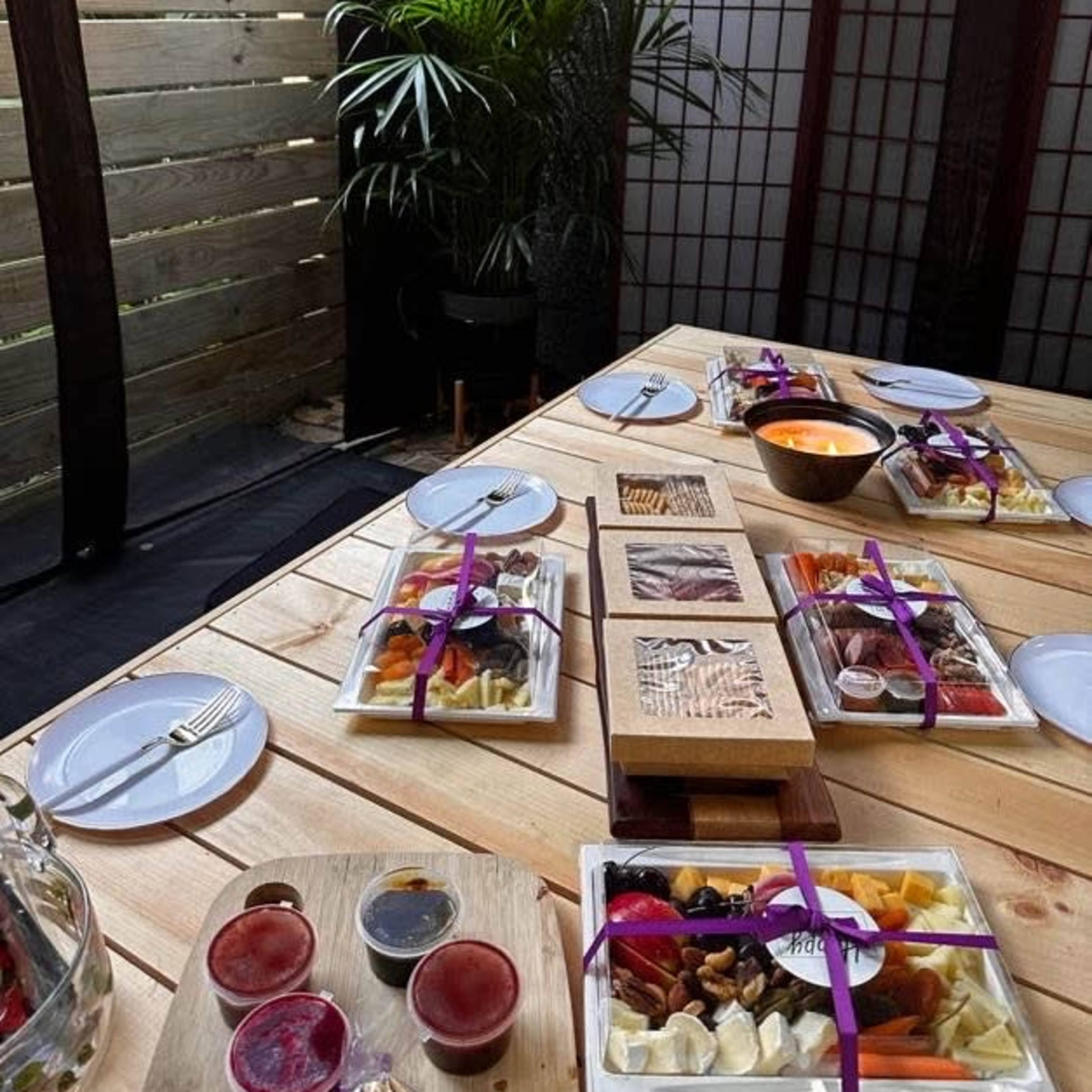 A wooden table is set outdoors with multiple plates of food, sauces, and a bowl, surrounded by partitions and greenery.
