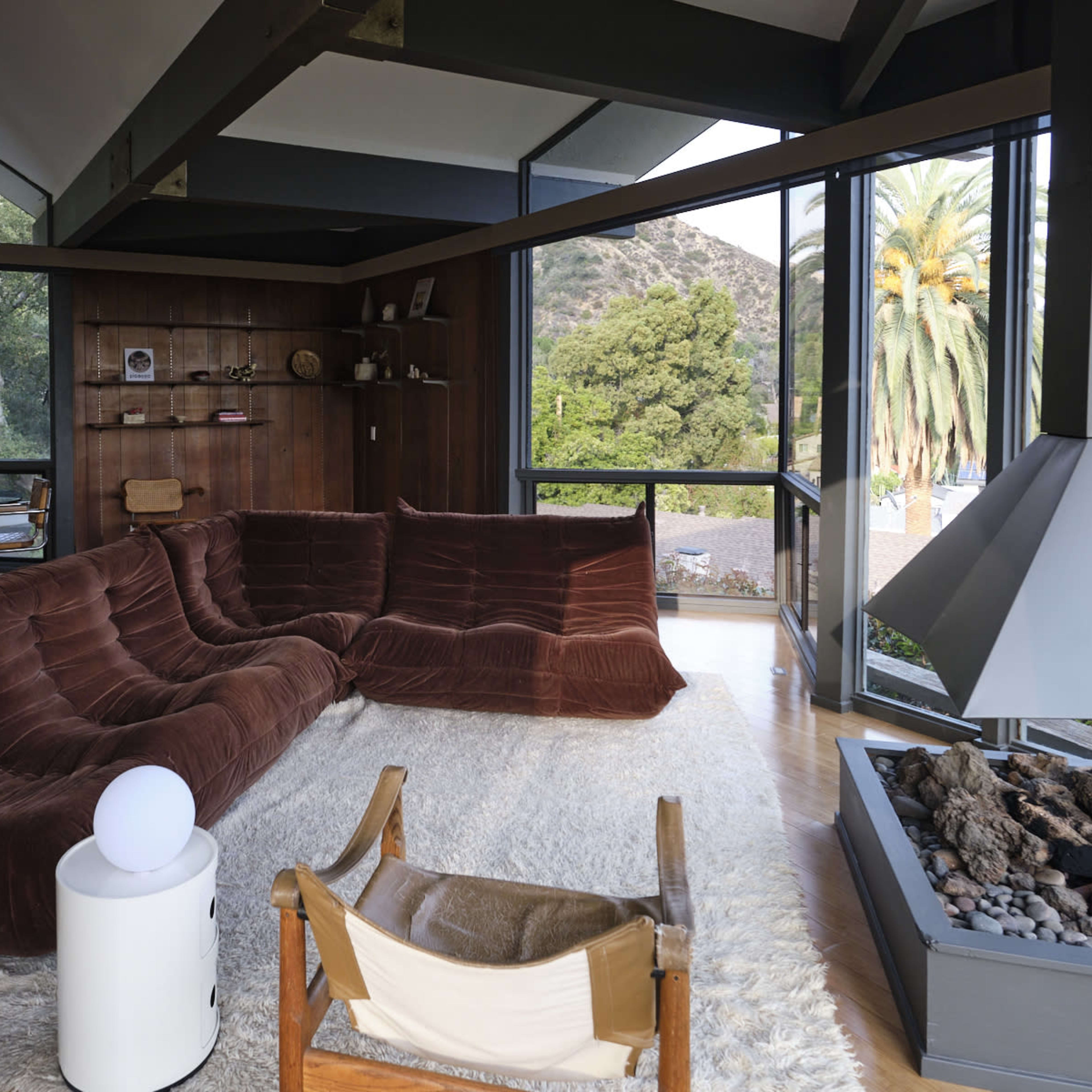 A modern living room with large windows, featuring a brown sectional sofa, a light-colored rug, a fireplace, and wooden accents.