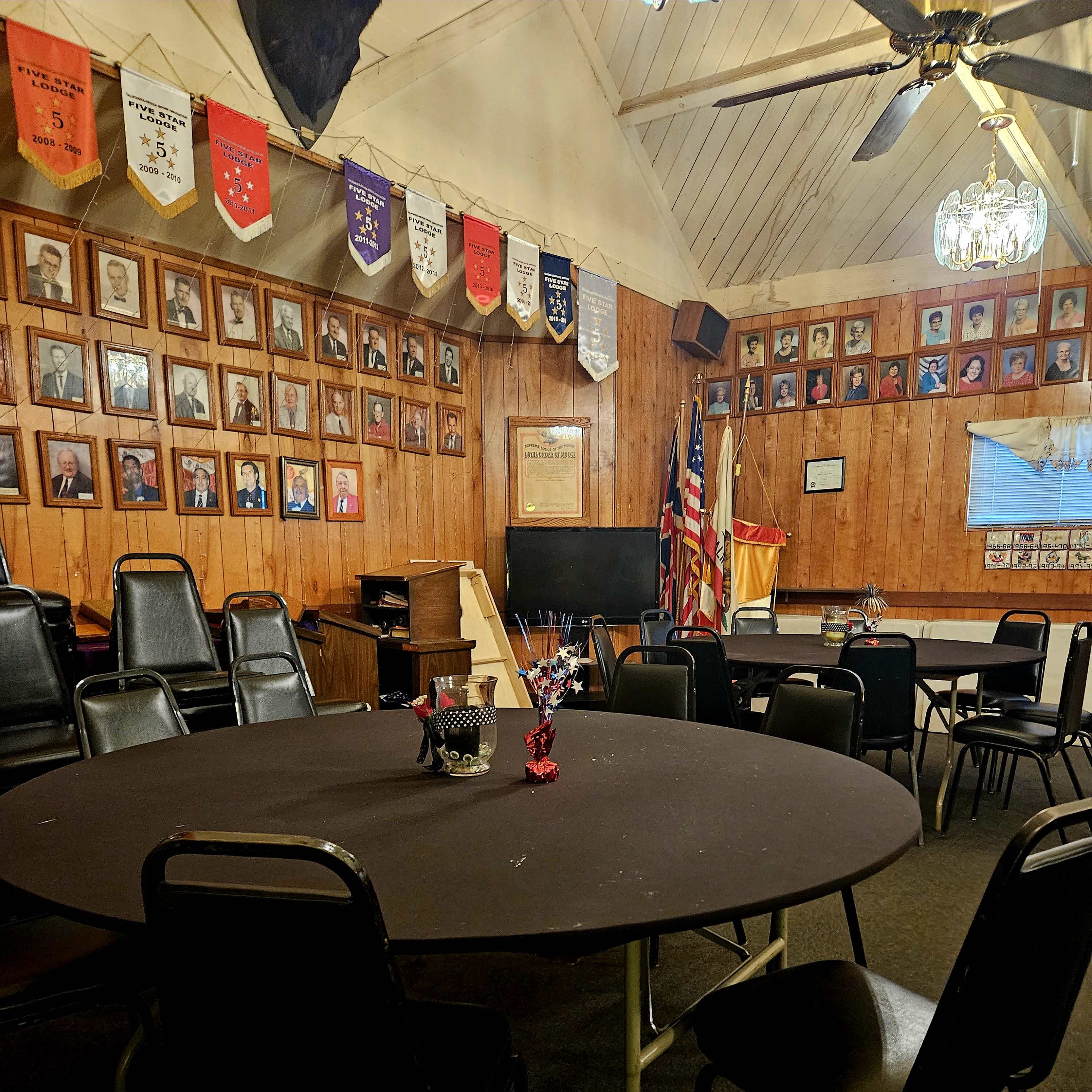 A meeting room with several round tables, black chairs, wooden walls adorned with framed photos and banners, and a projector screen at one end.