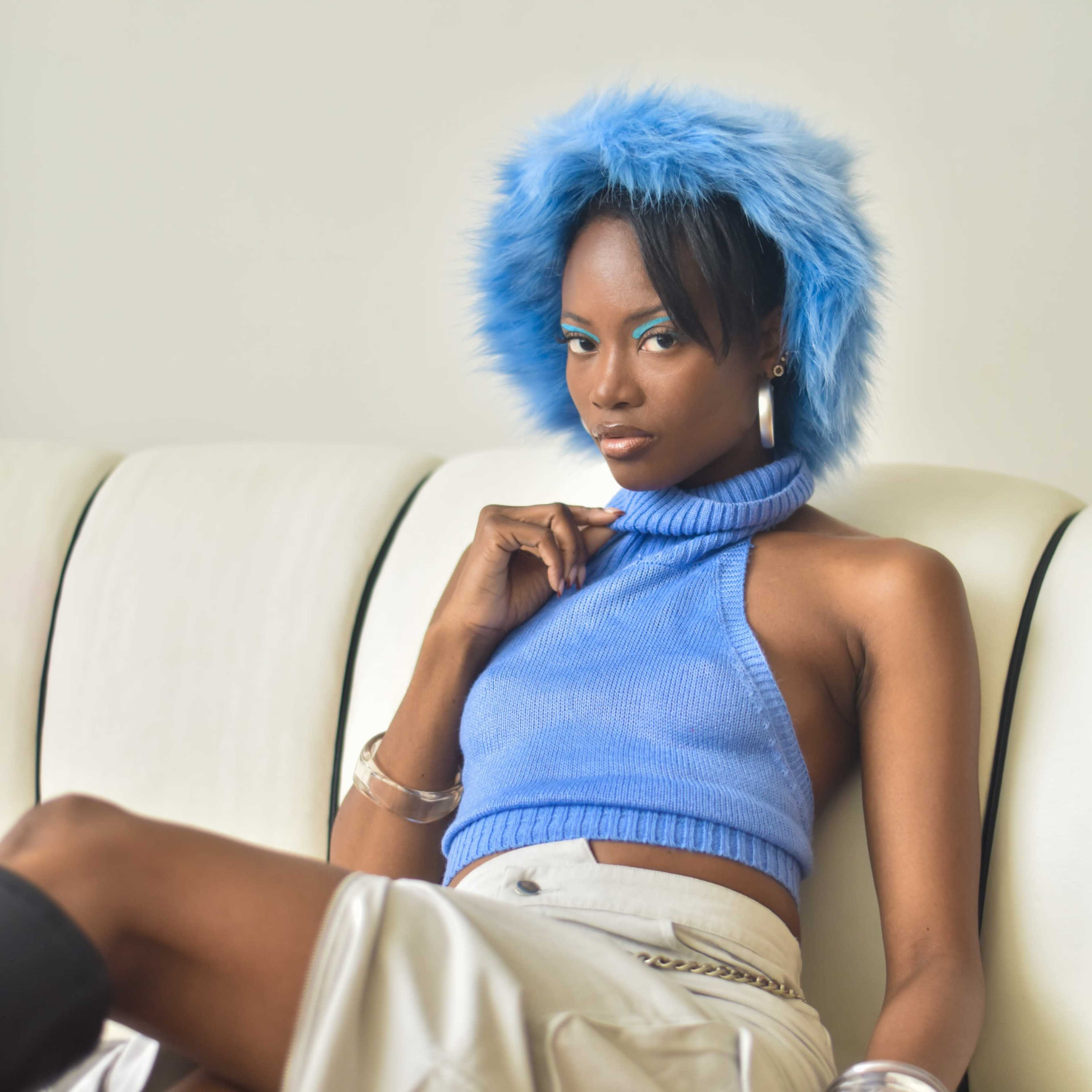 A person wearing a blue cropped sweater and a fluffy blue hat poses while seated on a white couch.
