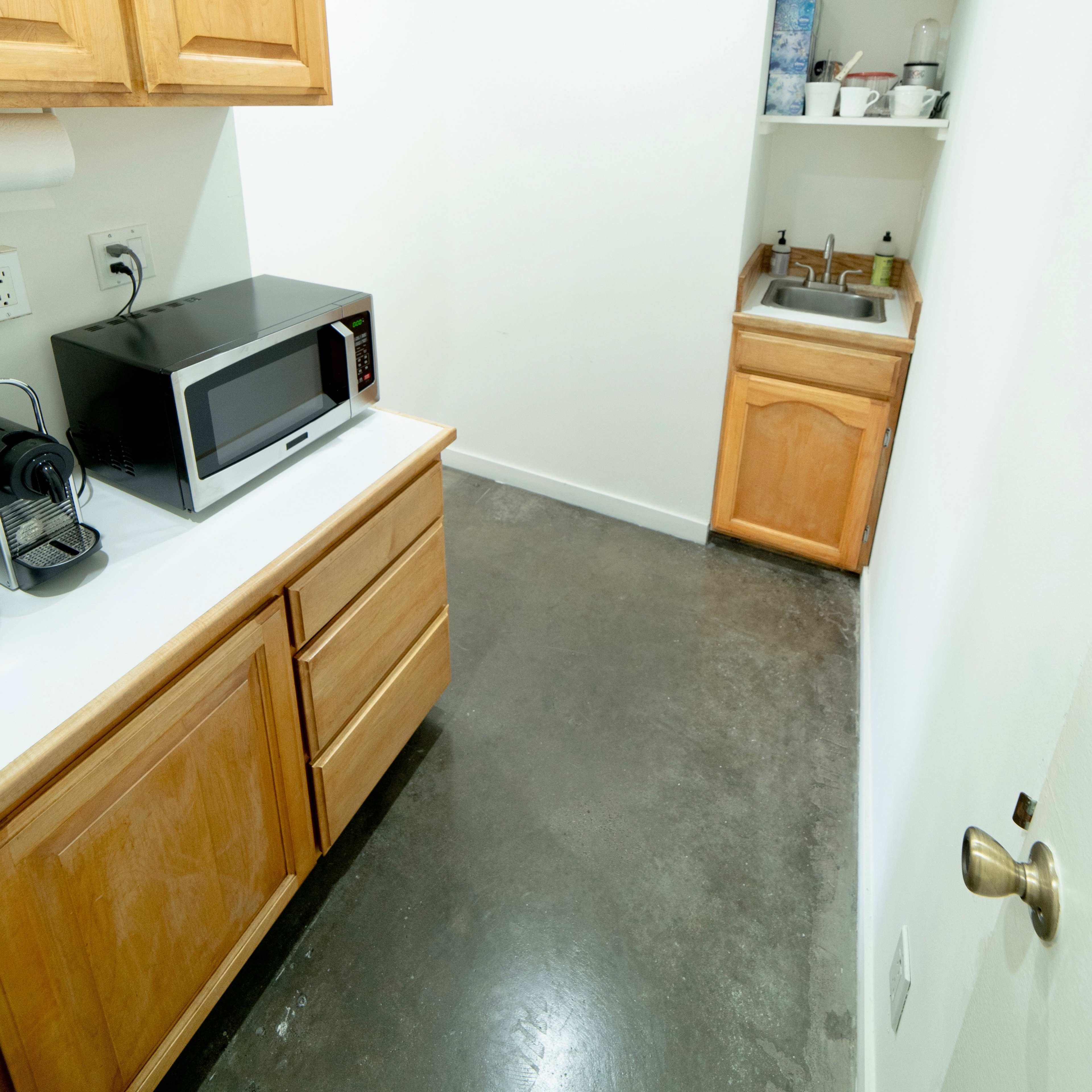 A narrow kitchen space featuring wooden cabinetry, a microwave, and a small sink against a white wall.