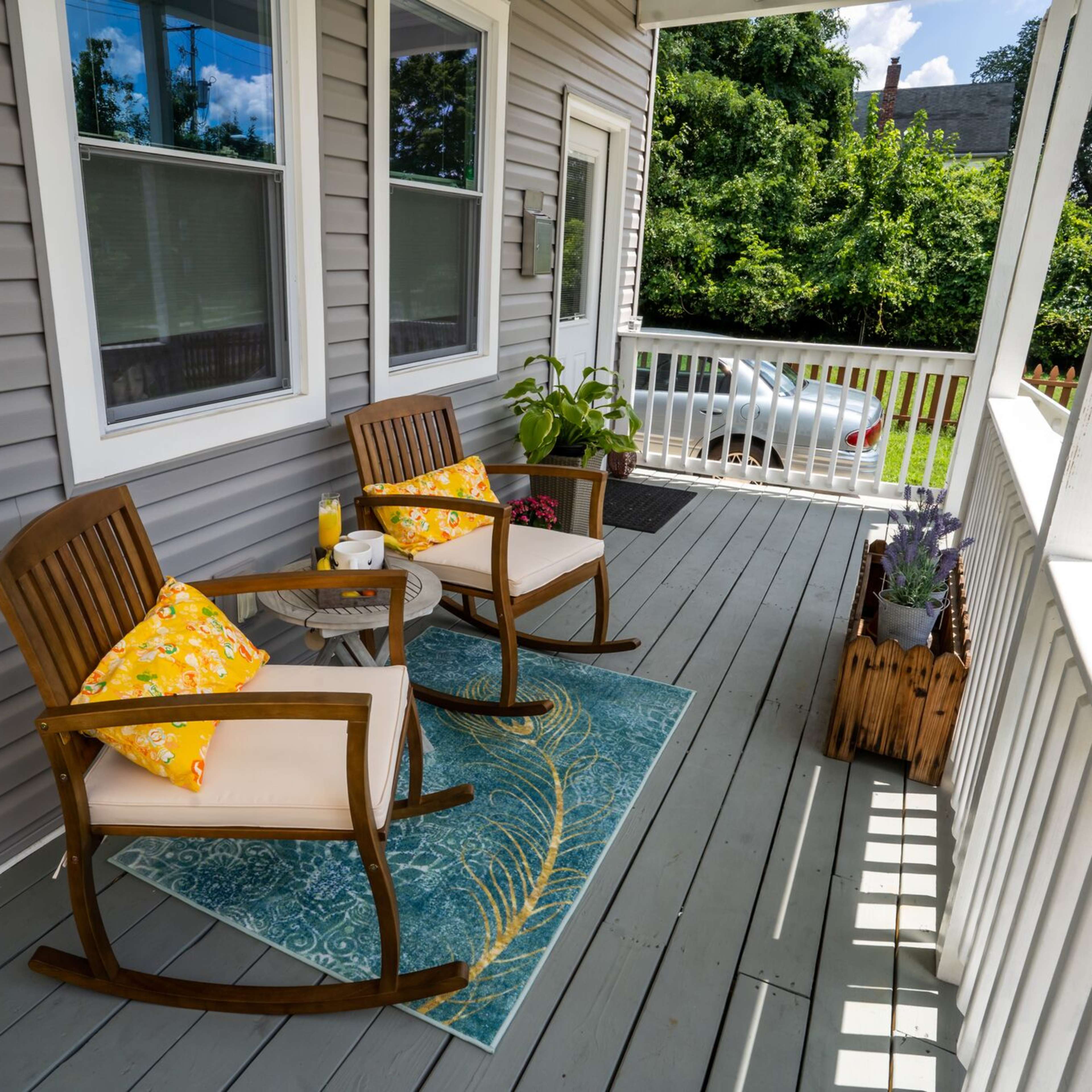 A porch with two wooden rocking chairs, a blue rug, and potted plants, set against a backdrop of a gray house.