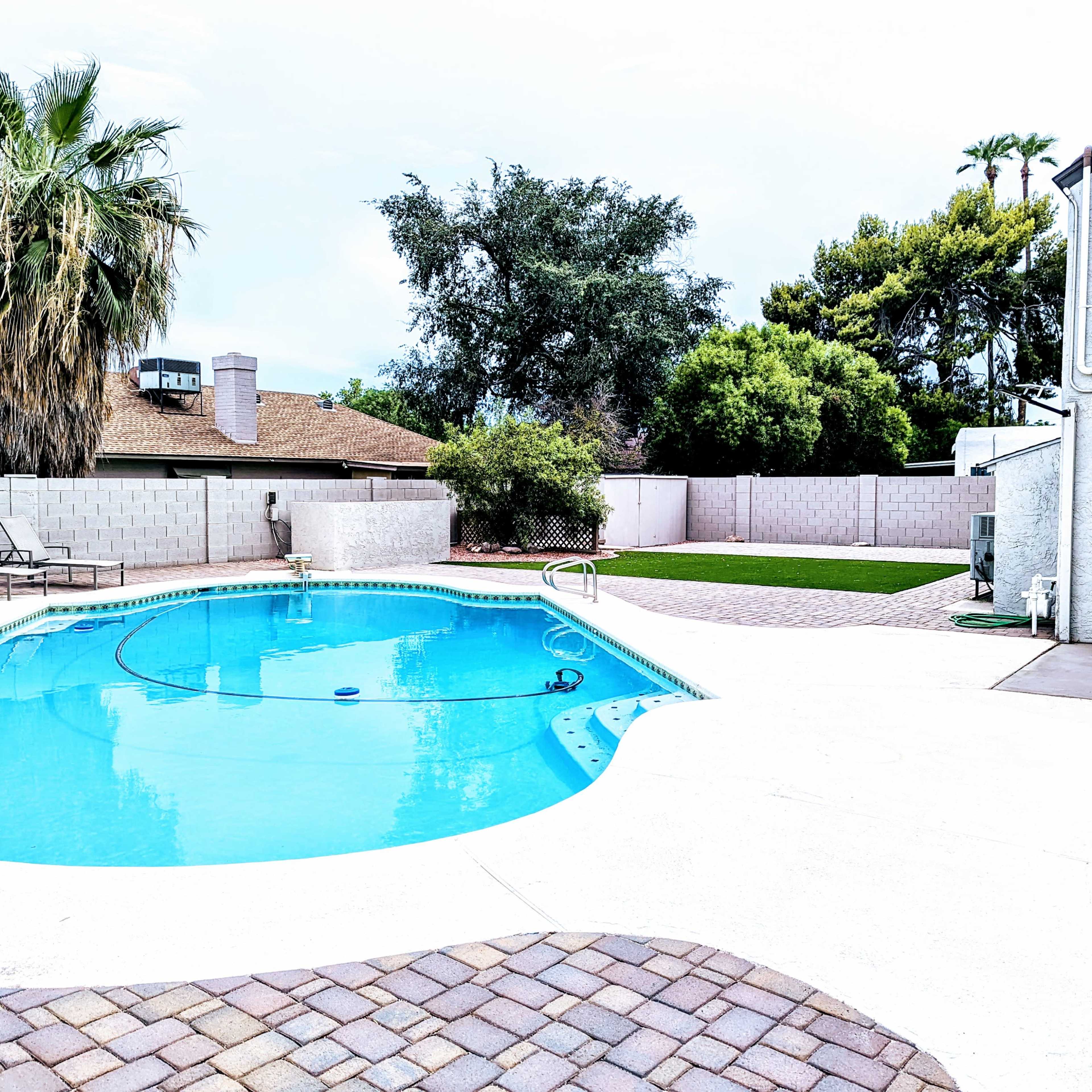 A swimming pool with a blue surface is surrounded by a paved deck and grass area in a residential backyard.