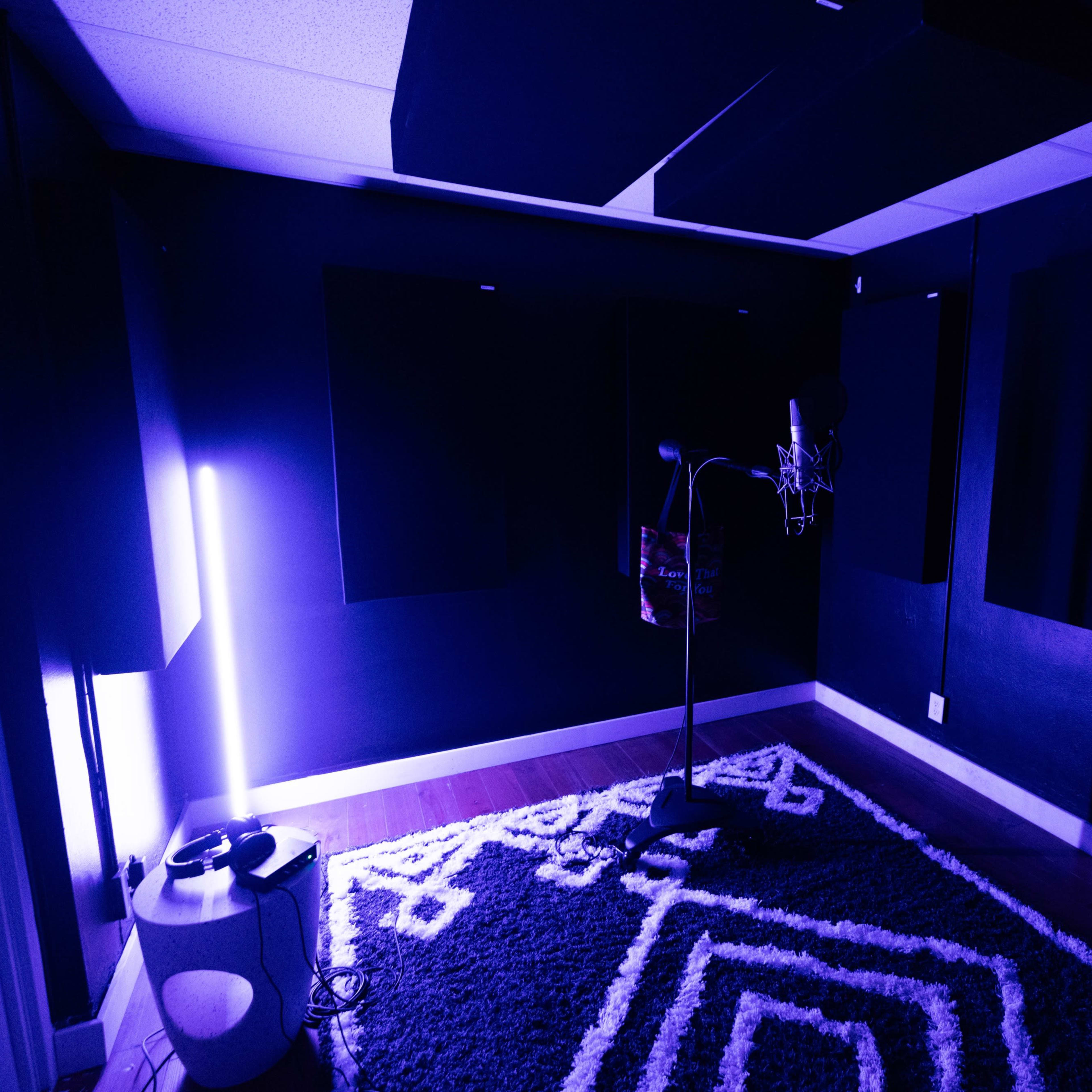 A small recording studio features a microphone on a stand, surrounded by soundproofing panels and illuminated by purple lighting.