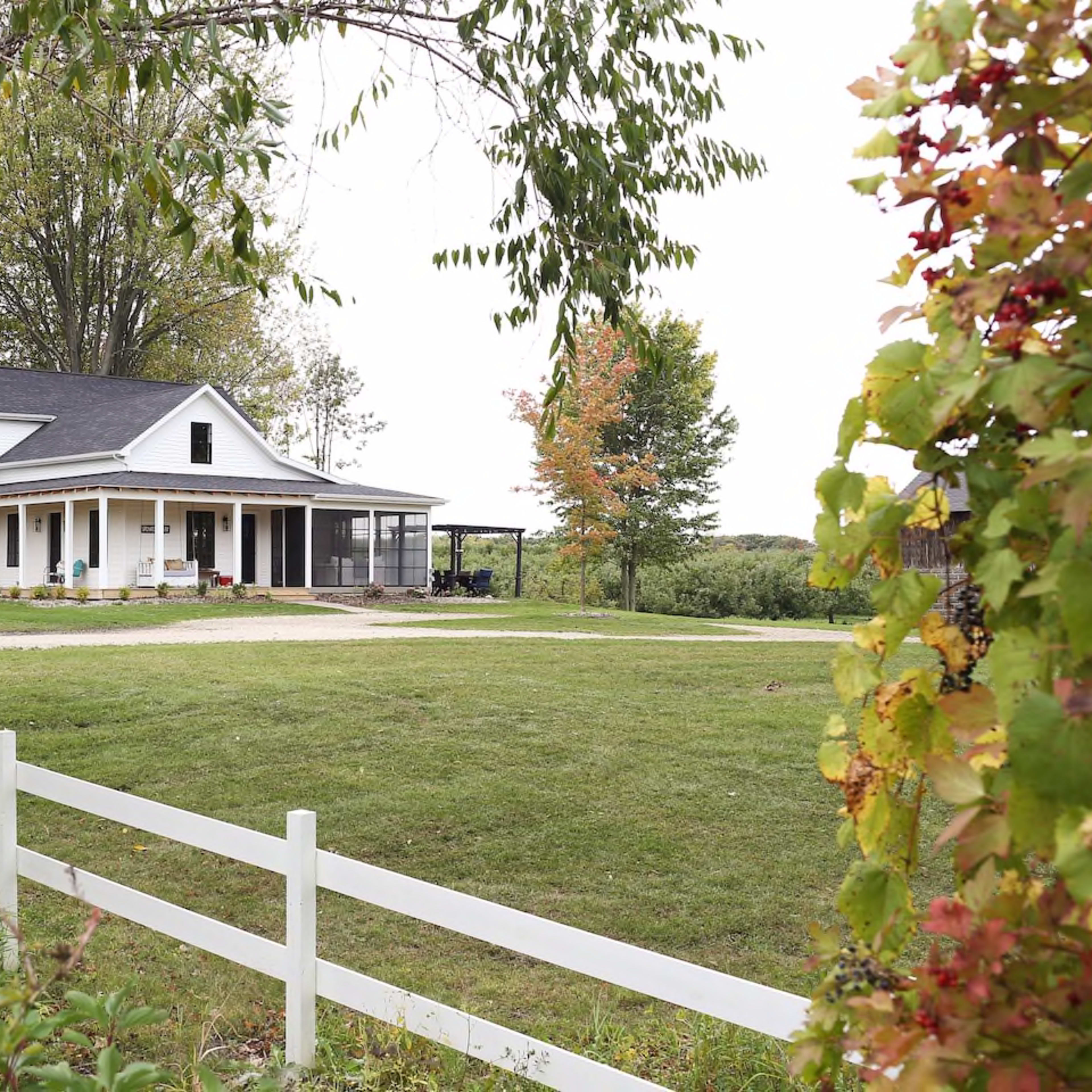 A white farmhouse with a black roof and front porch sits on a grassy lawn surrounded by trees and a white fence.