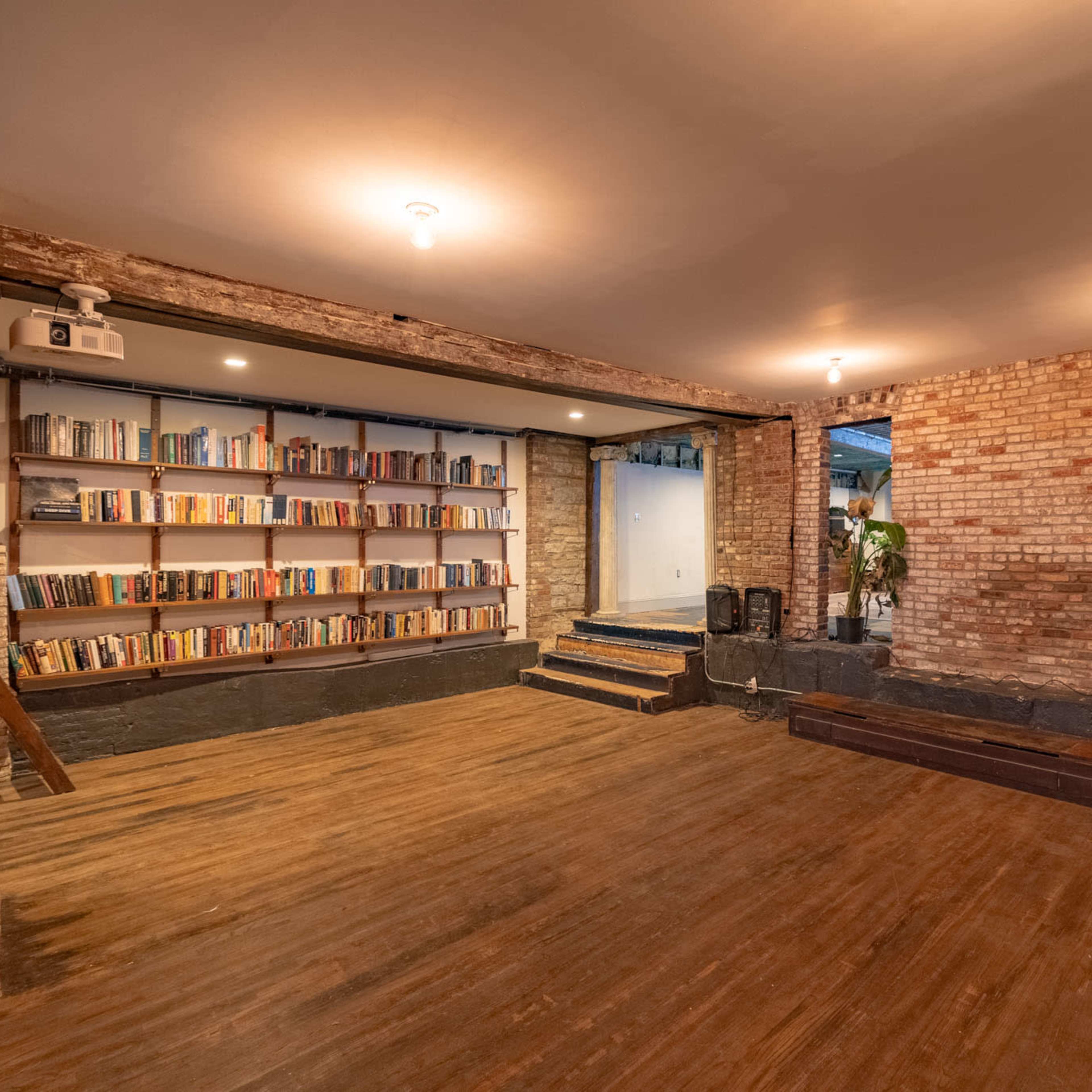 An interior space with exposed brick walls, a wooden floor, and a bookshelf filled with books, leading to a set of steps at the back.