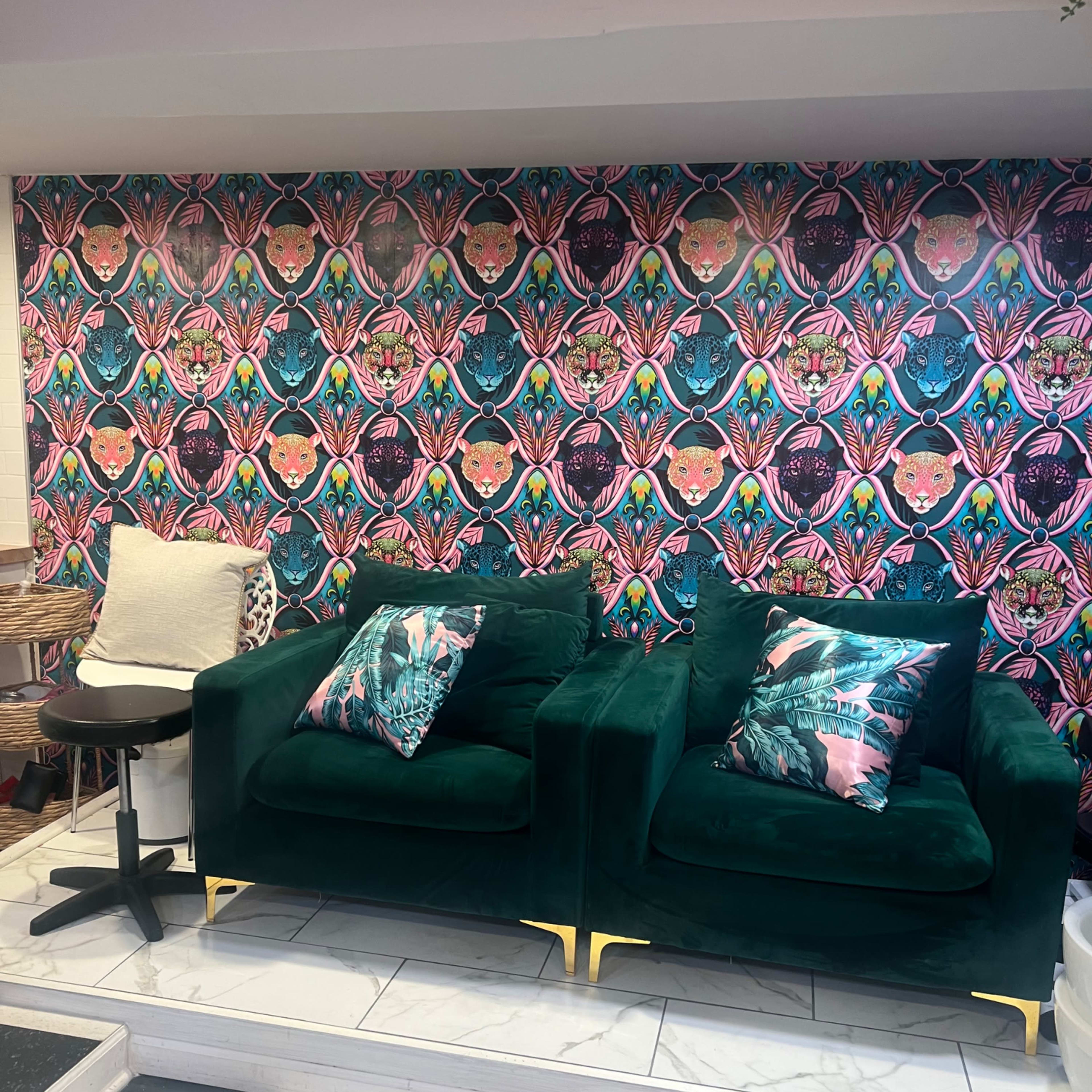 Two green velvet chairs with decorative pillows are positioned against a colorful patterned wallpaper in a stylish interior space.