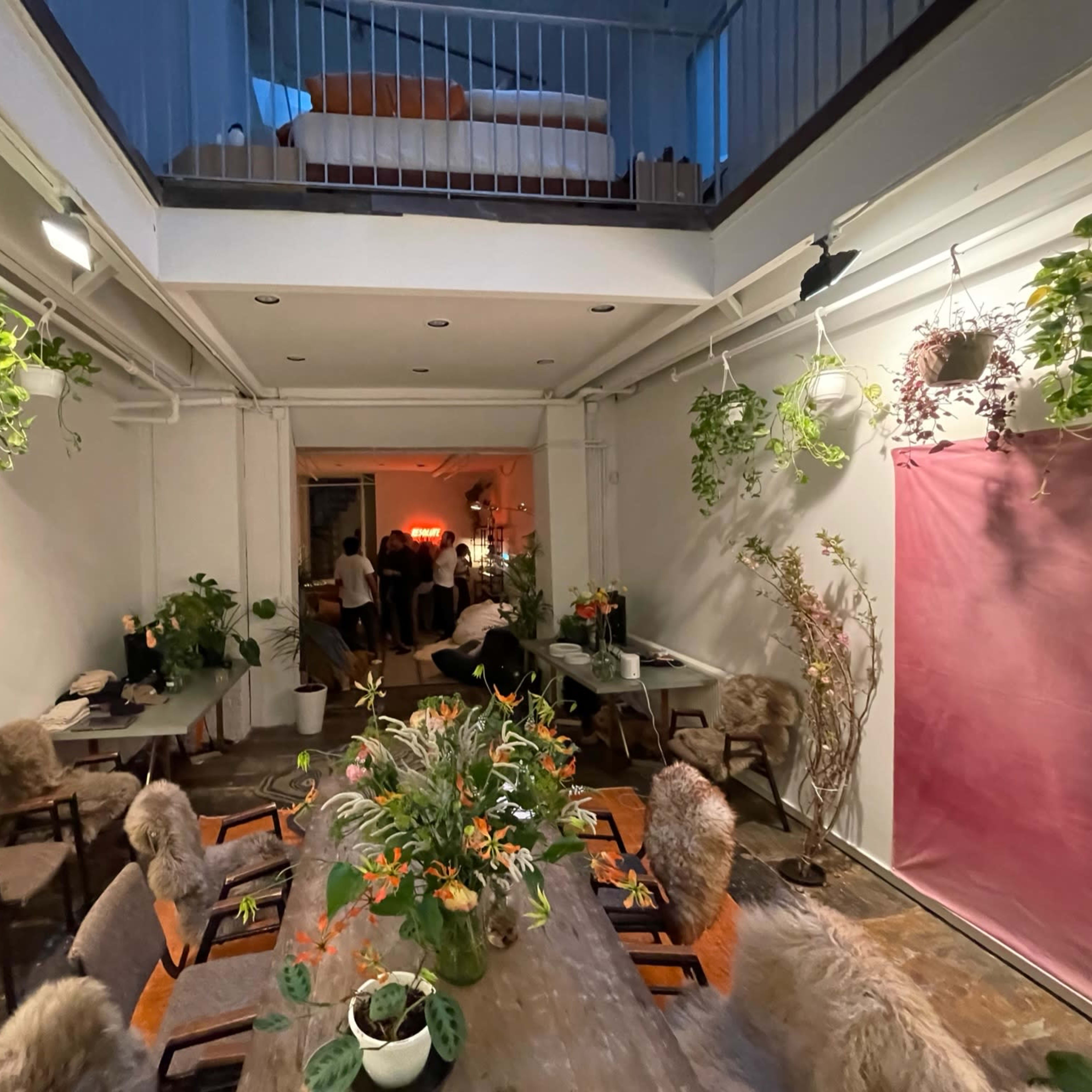 A well-decorated indoor space with a long dining table surrounded by chairs, plants hanging from the ceiling, and a visible upper level with a bed.