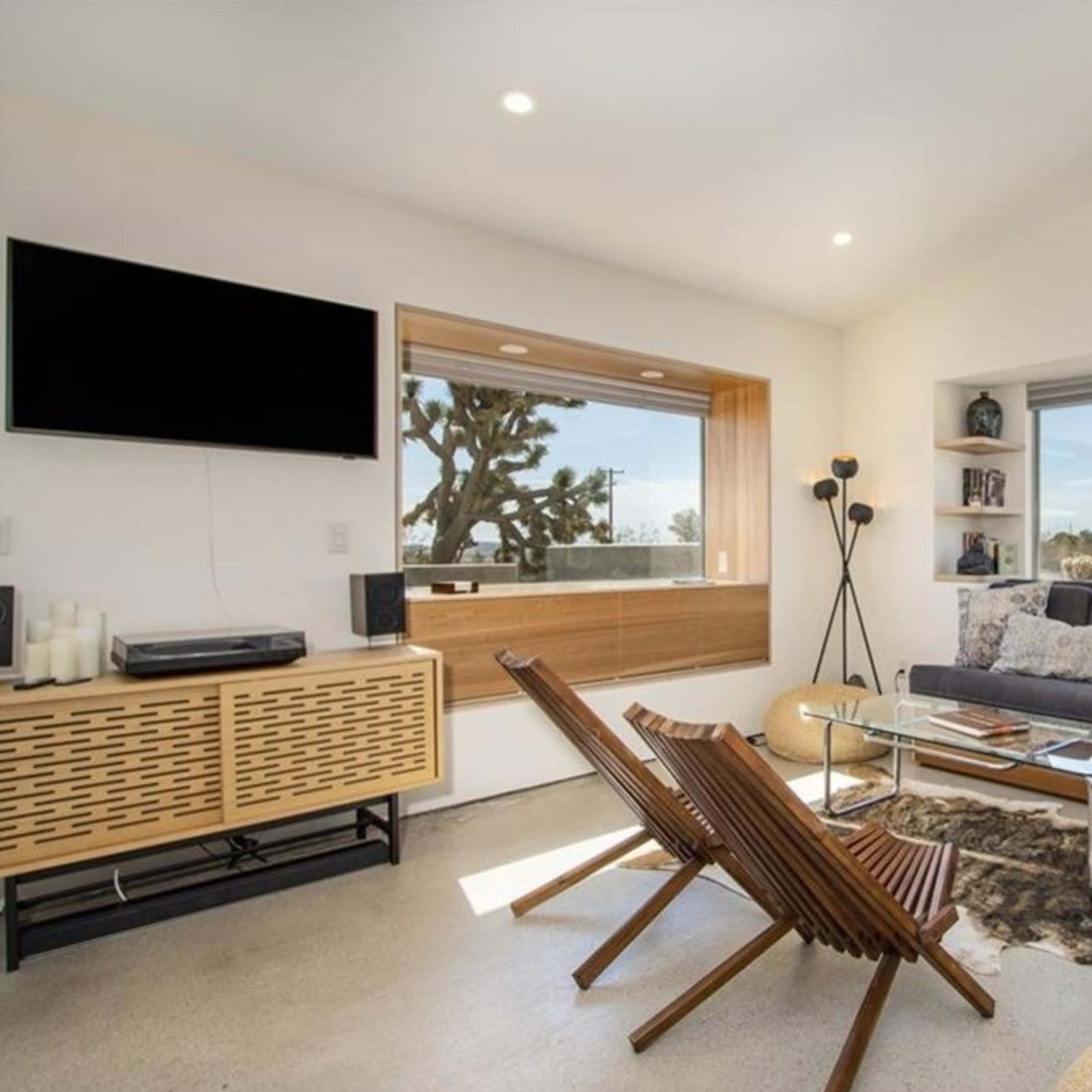 A modern living room with a television mounted on the wall, a large window providing natural light, and a mix of wooden furniture and soft furnishings.