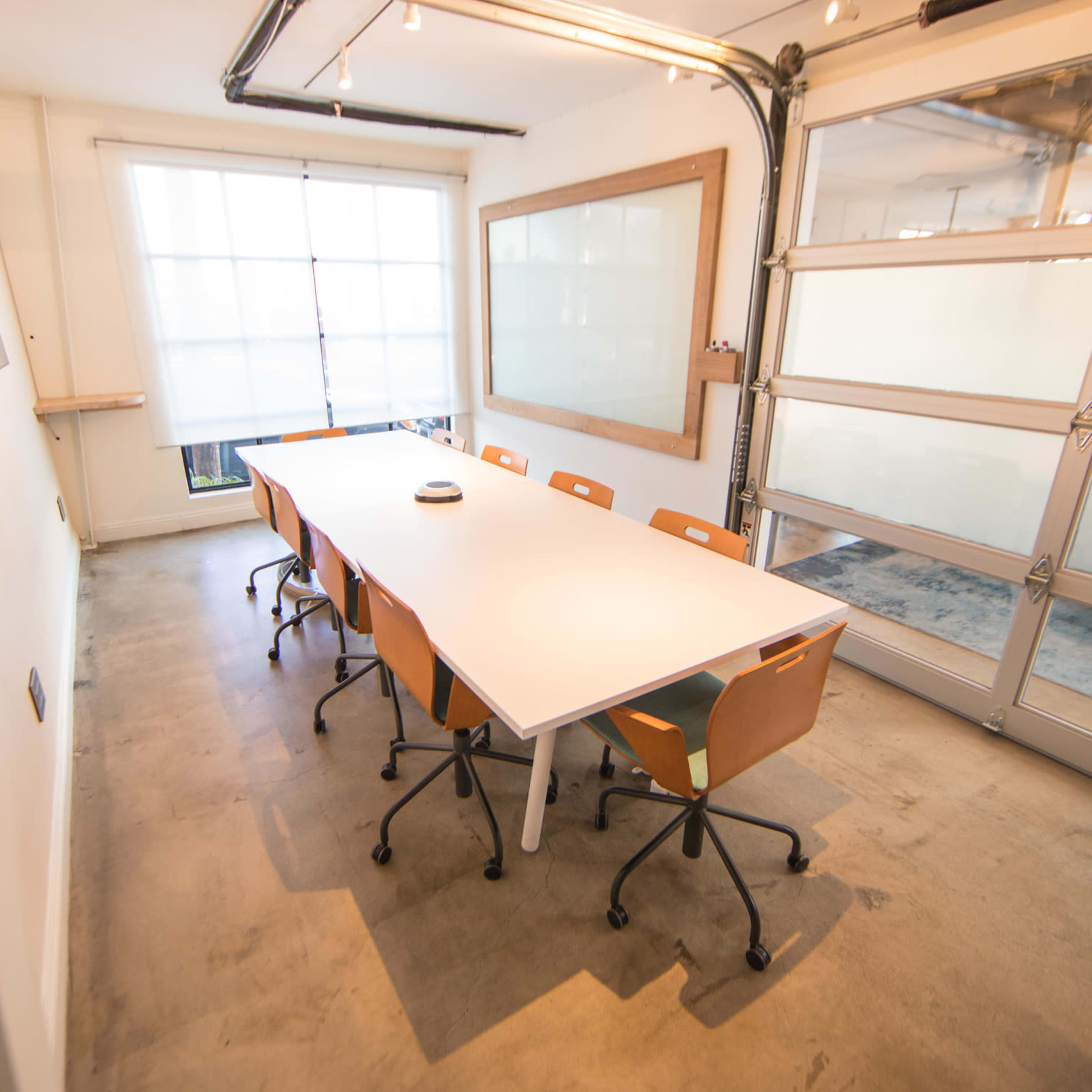 A modern conference room with a long white table surrounded by several orange chairs, large windows, and a glass garage door.