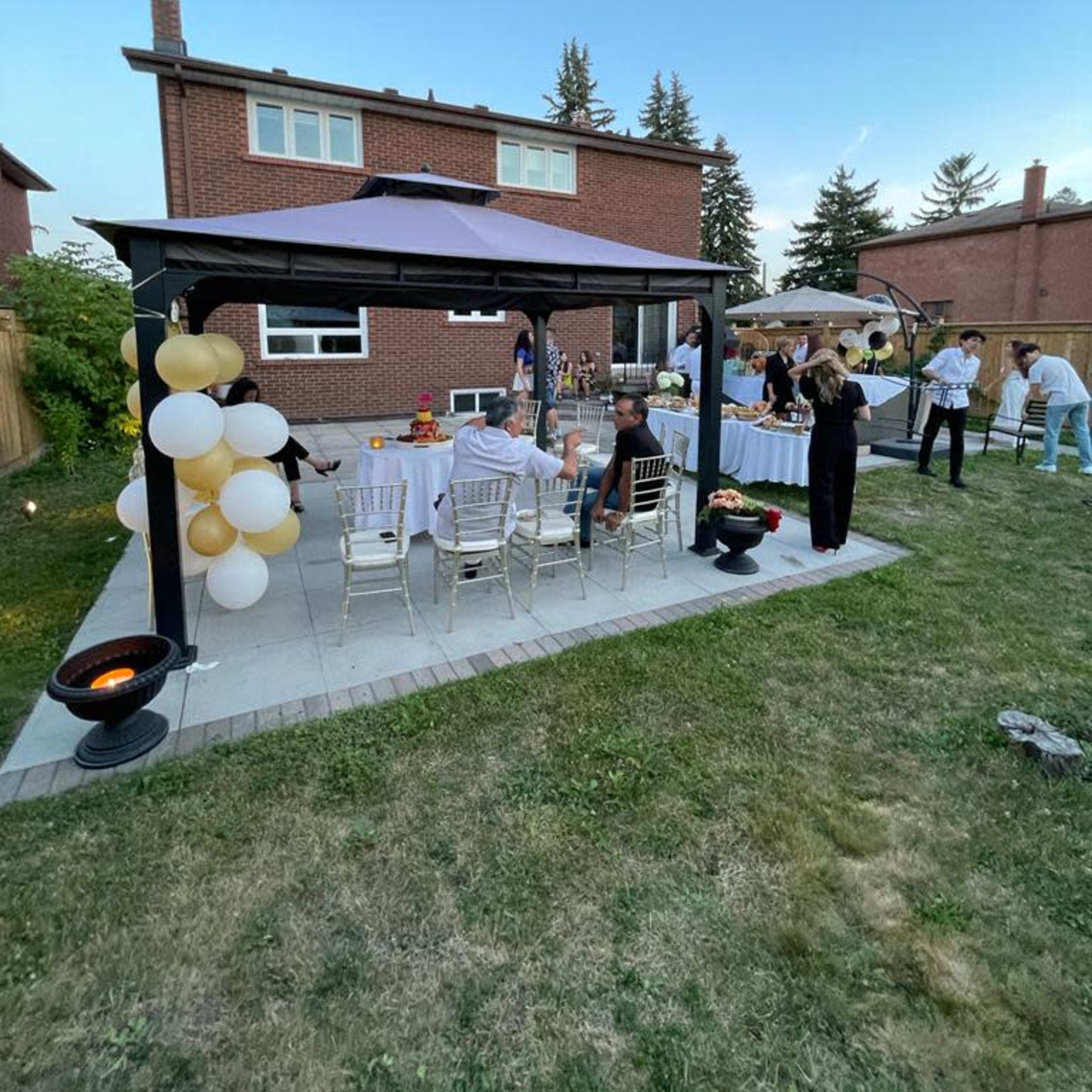 A group of people is gathered under a gazebo in a backyard, with a table set for an outdoor event decorated with balloons.