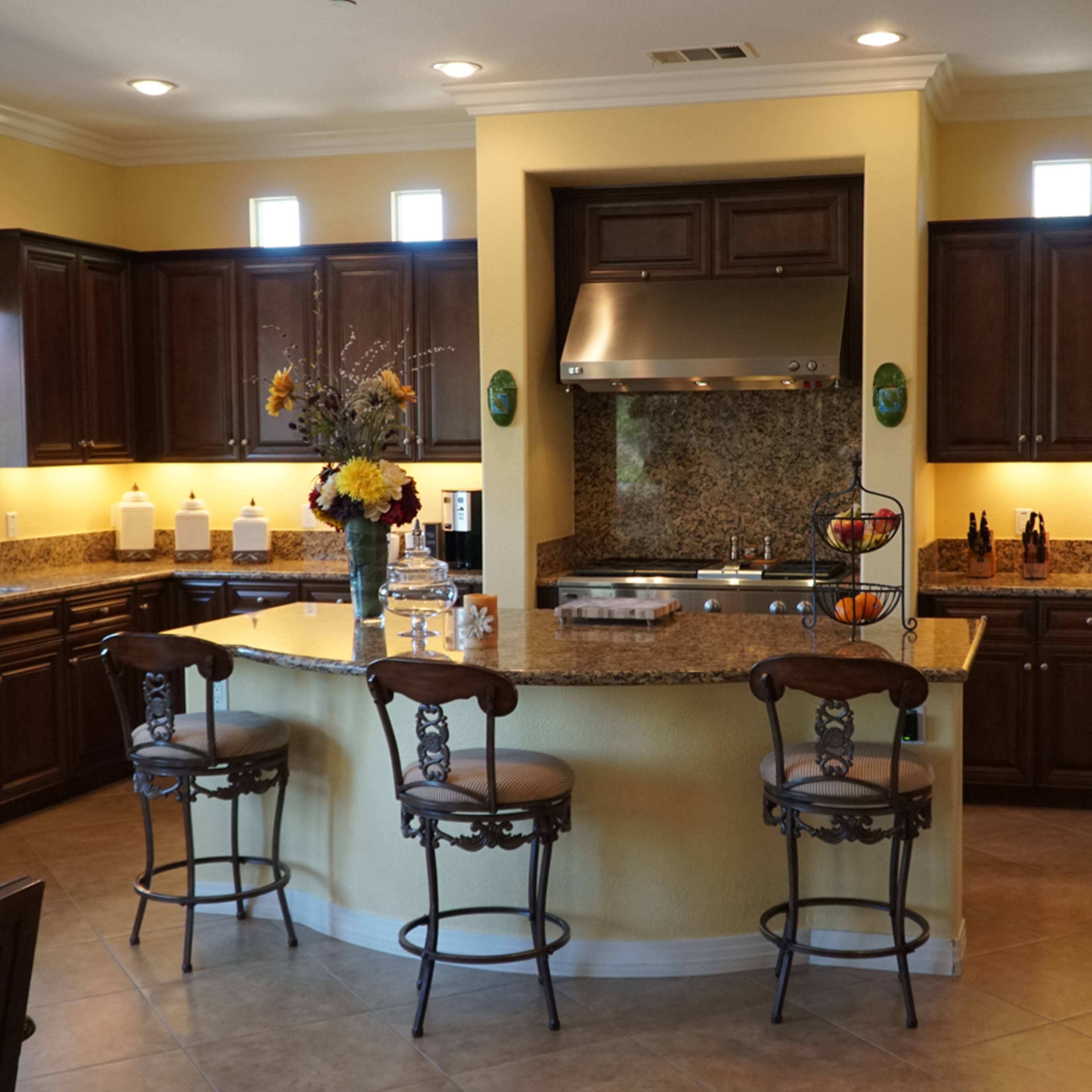 A modern kitchen featuring wooden cabinets, granite countertops, and a central island with three bar stools.