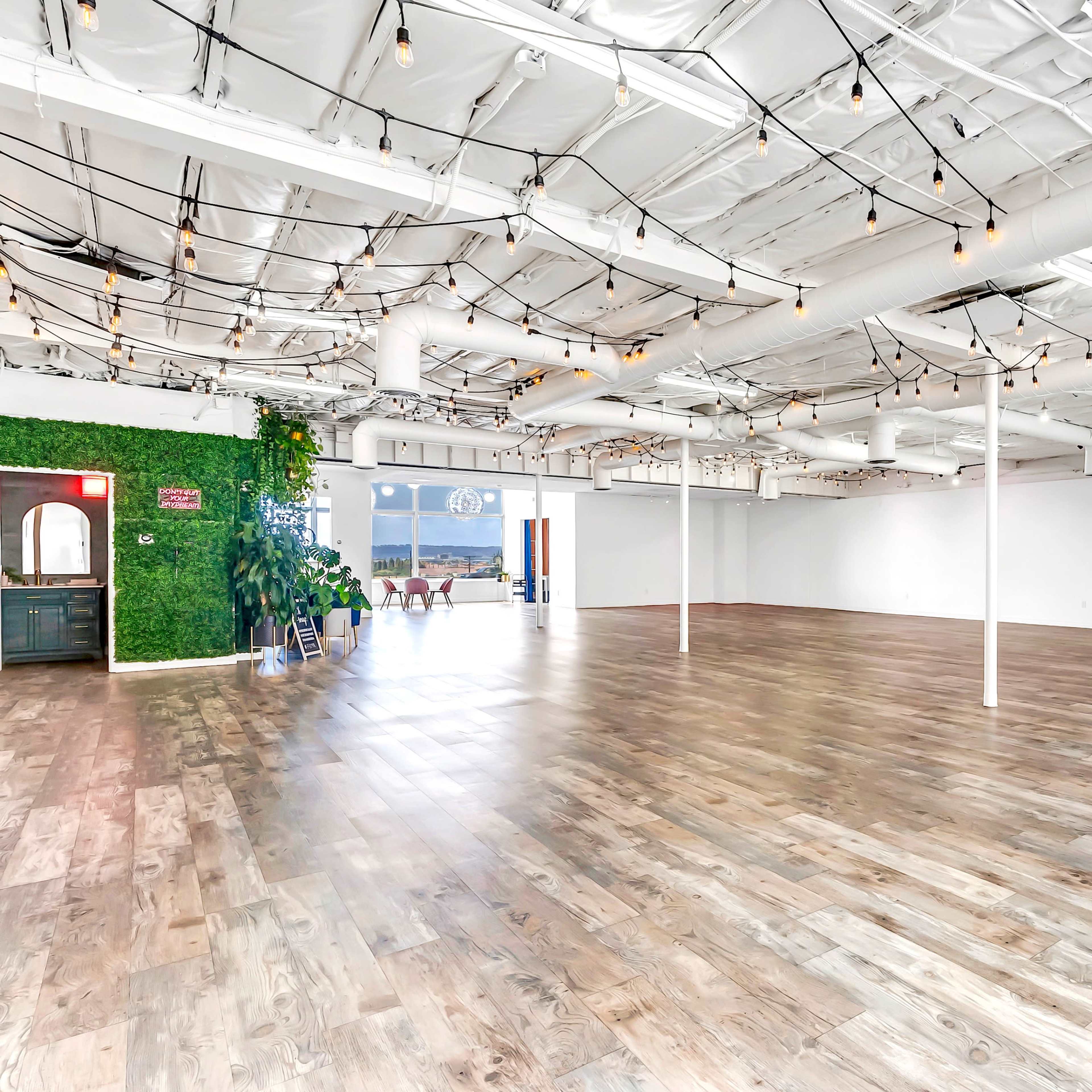 A spacious, empty room with wooden floors, white walls, and exposed ceiling beams adorned with hanging string lights.