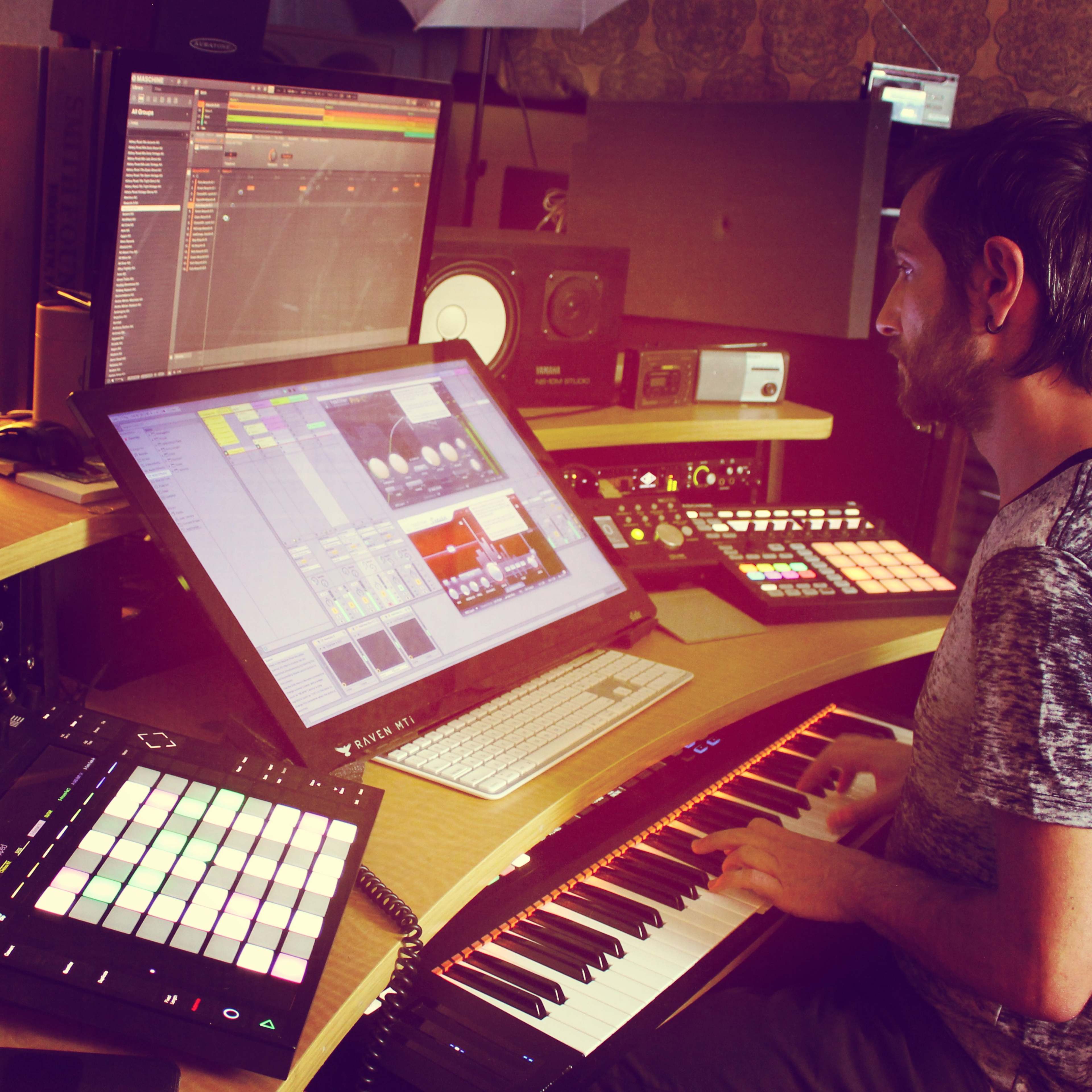 A person plays a keyboard while working on music production software at a computer workstation surrounded by electronic equipment.