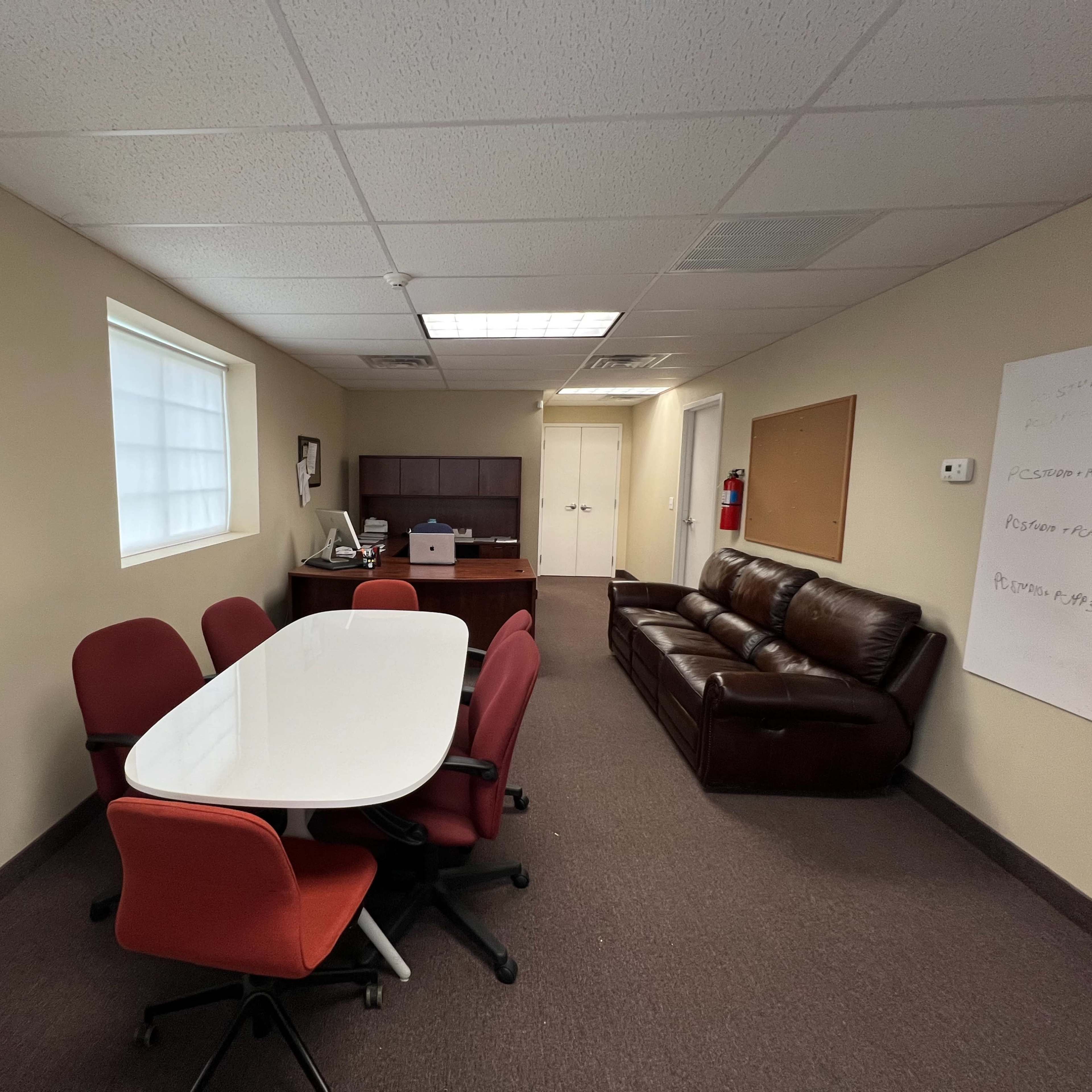 A small office space with a conference table surrounded by red chairs, a brown leather couch, and a desk against the wall.