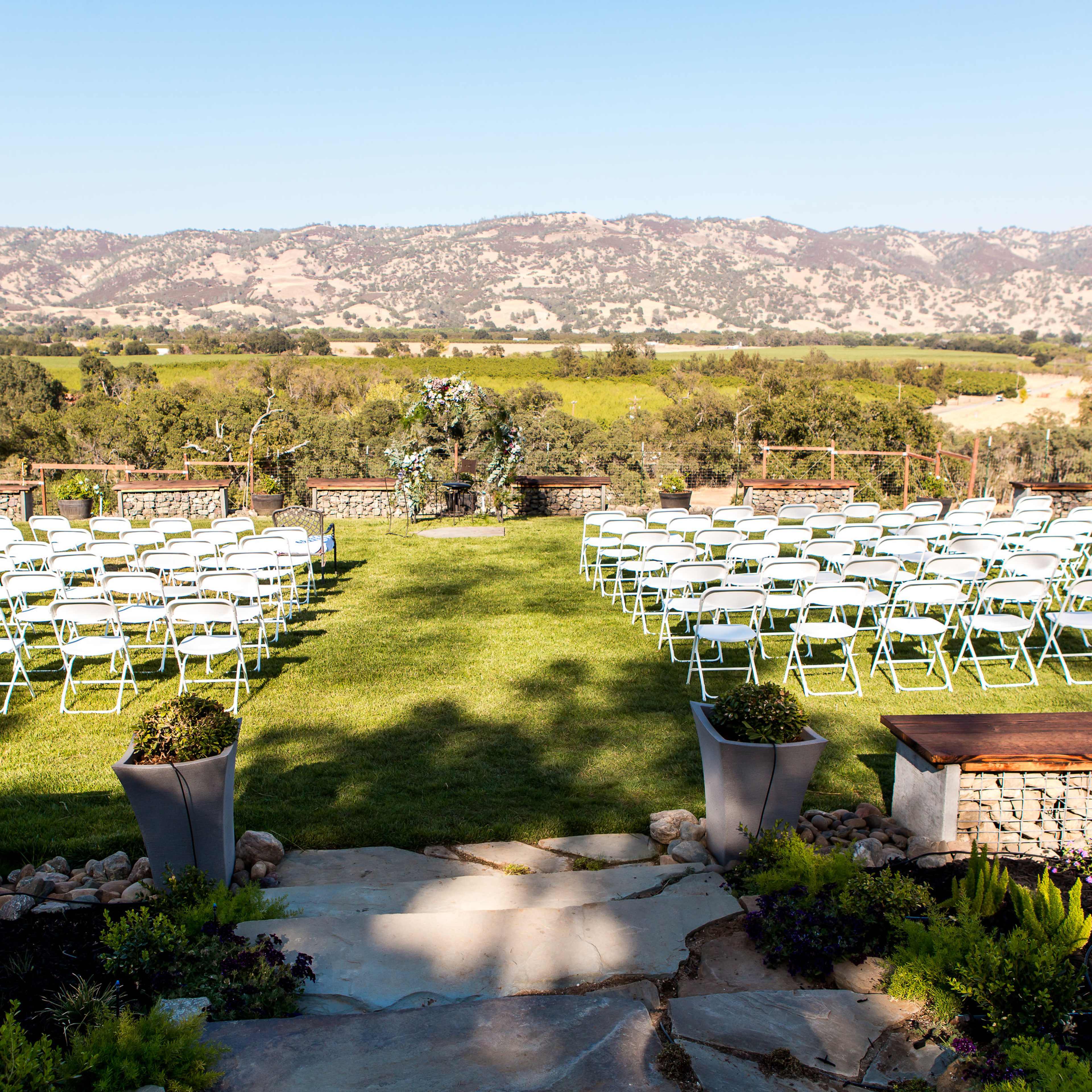 A wedding ceremony setup with rows of white chairs arranged on a grassy lawn overlooking a mountainous landscape.