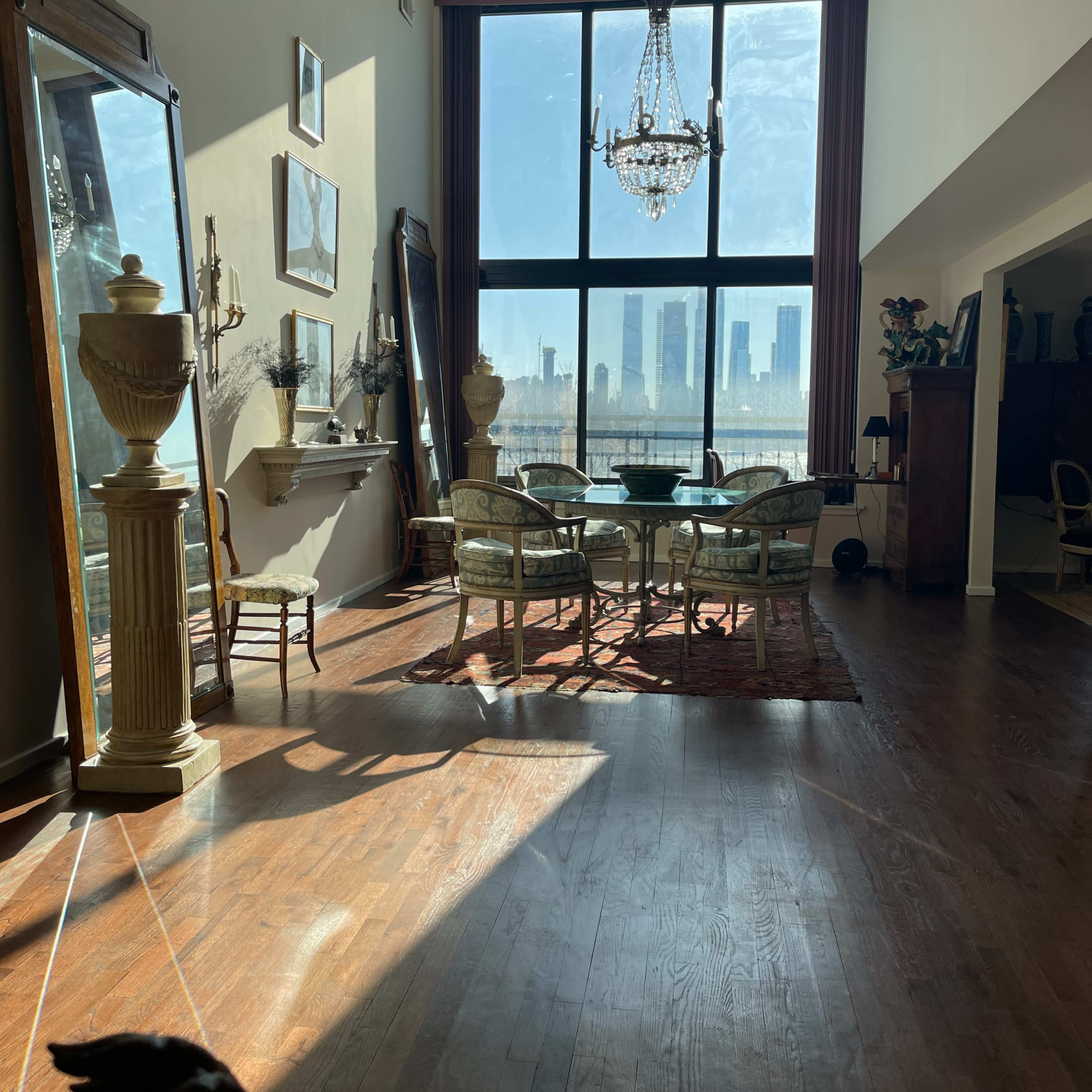 A sunlit dining area features a large window with a city skyline view, a chandelier, and ornate furniture.