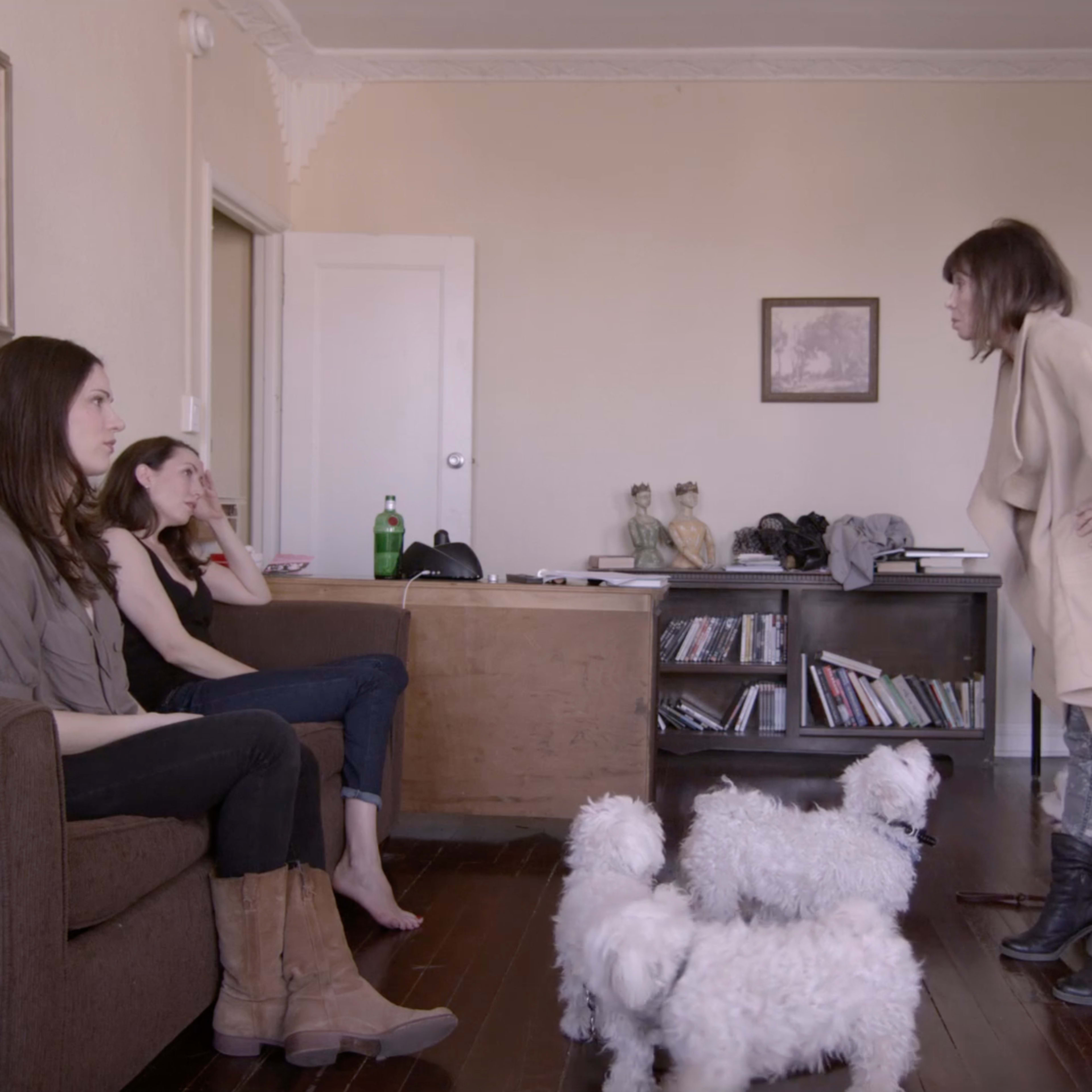 Two women sit on a couch while a third woman stands near them, engaging in conversation as three small white dogs gather on the floor.