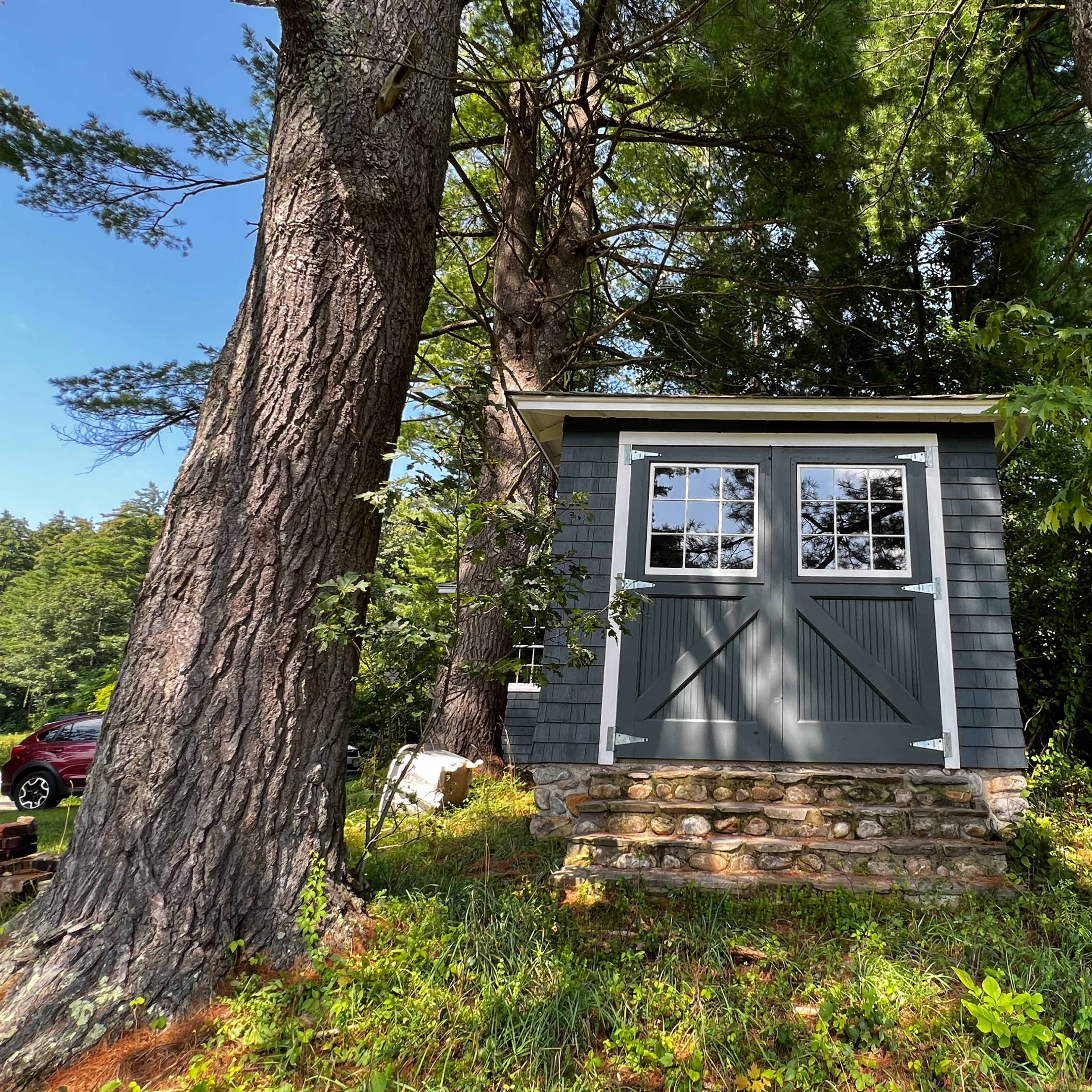 A small, gray shed with double doors is situated on a stone foundation, surrounded by tall trees and greenery.