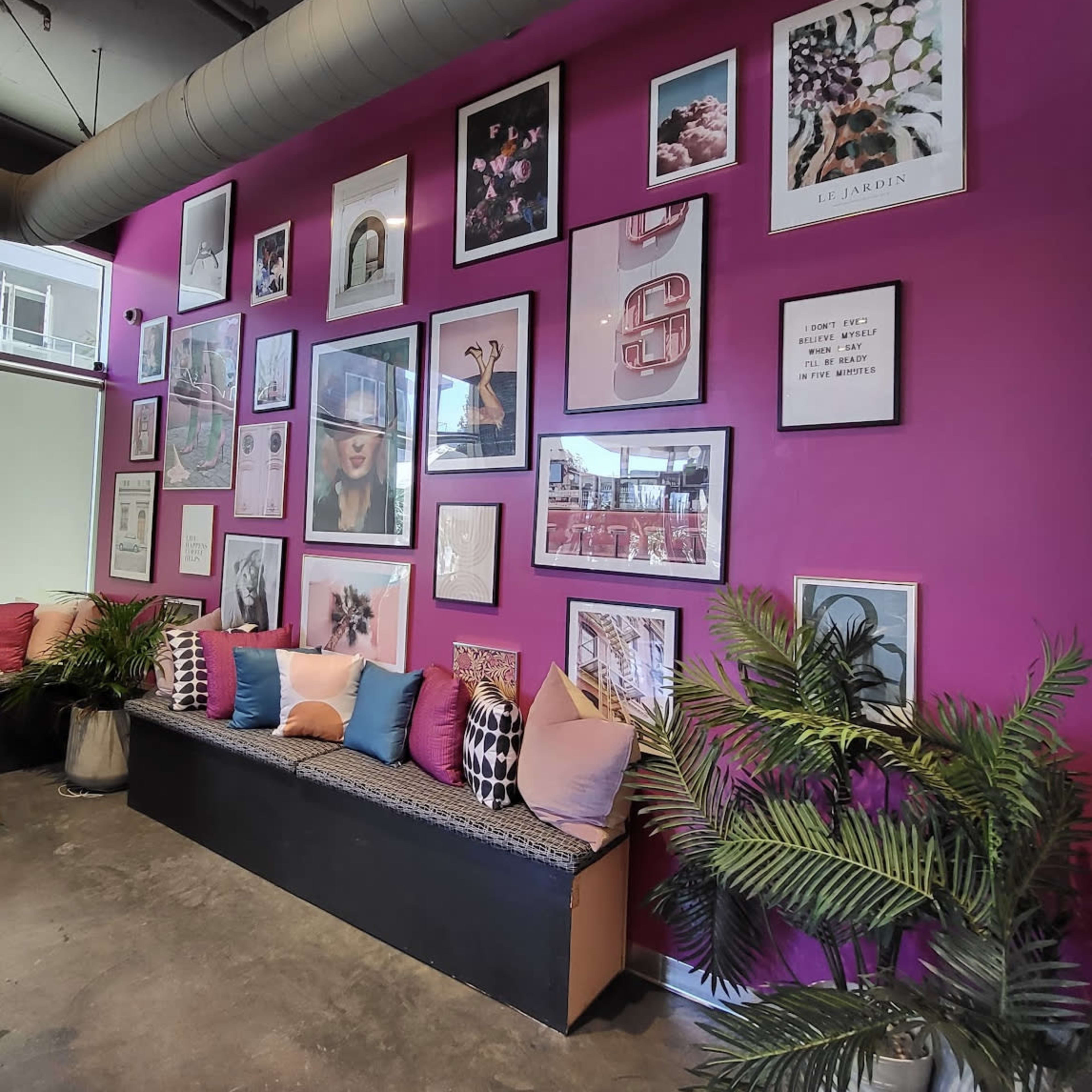 The scene features a wall painted pink, adorned with various framed artworks and photographs, alongside a seating area with colorful cushions and potted plants.